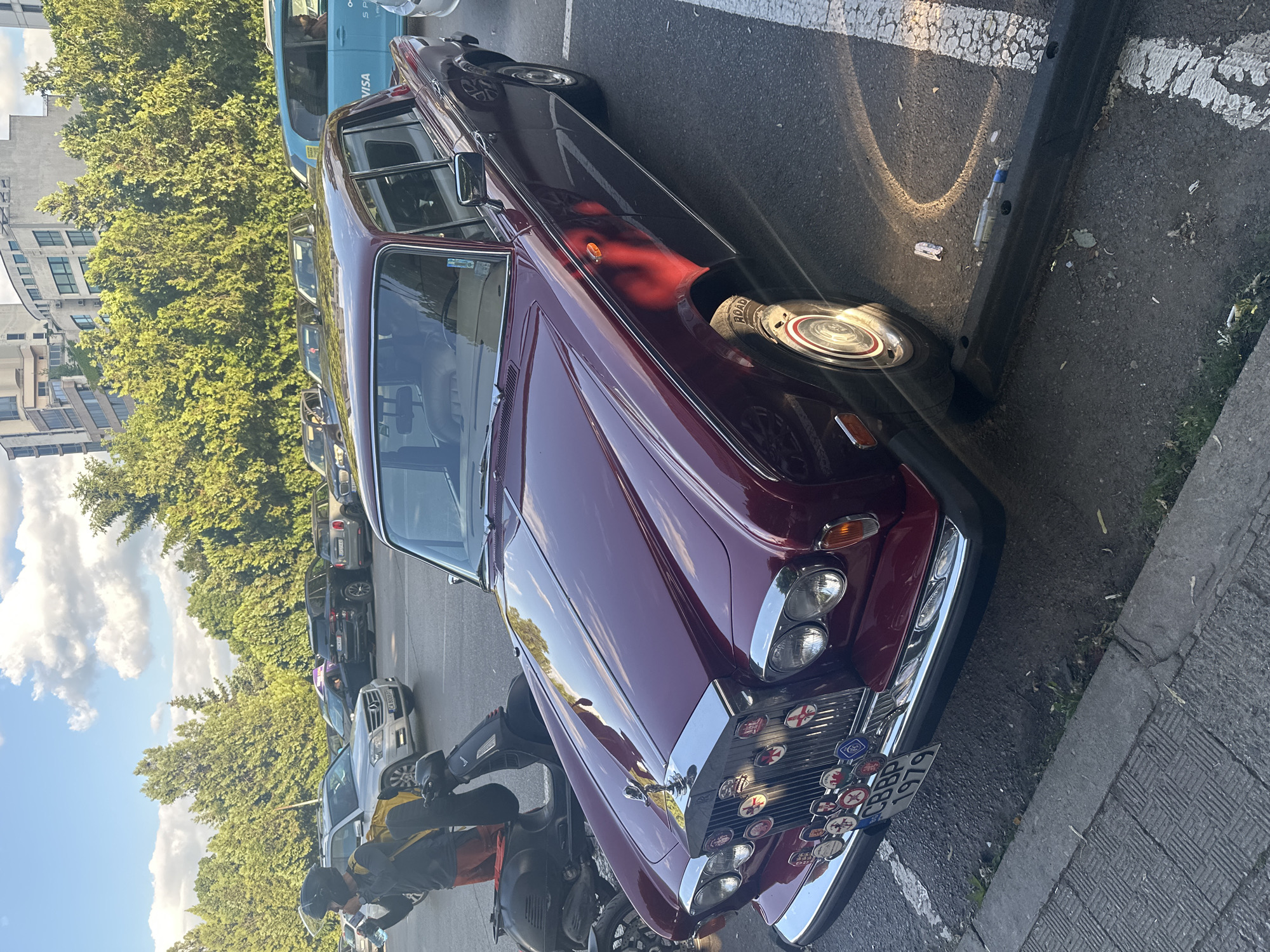 The image shows a maroon Rolls-Royce parked on a street in Sofia, Bulgaria. The car has a license plate that reads "CBBP 1979". A motorcyclist in a black jacket and helmet stands next to the Rolls-Royce. Other parked vehicles, trees, and buildings are visible in the background, along with a blue sky with scattered clouds. The scene appears to be in an urban environment. The Rolls-Royce is adorned with several circular badges on its front grille. The parking area is delineated with white lines. There is a black parking barrier on the right side of the image.