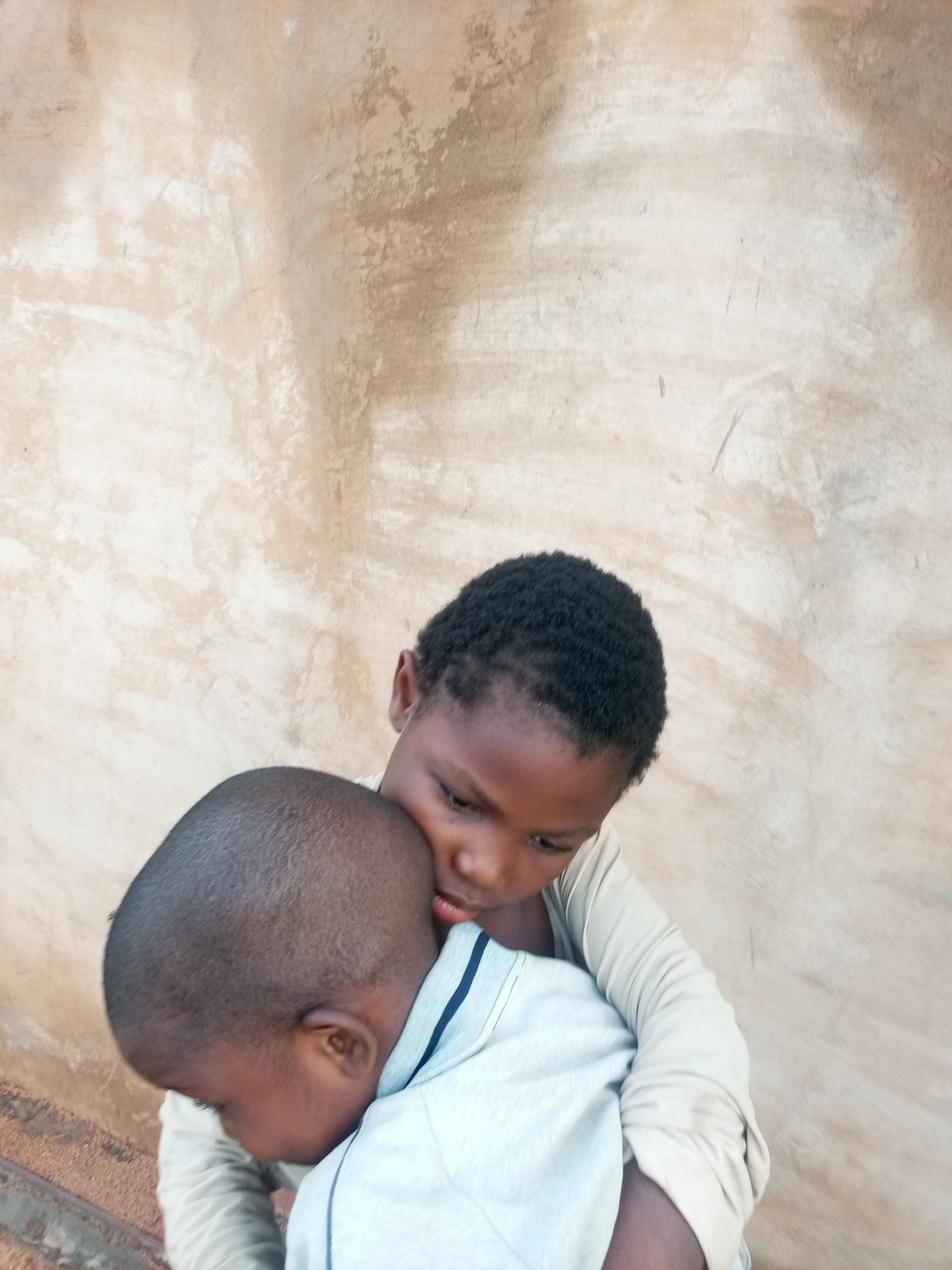 The image shows two young children embracing against a textured, light-brown wall. One child, facing away from the camera, is being held by the other child, whose head is resting on the first child's shoulder, with their eyes downcast. Both children appear to be wearing light-colored shirts. The background wall exhibits variations in tone and texture, suggesting an outdoor or unfinished interior setting. The lighting is soft, casting subtle shadows and highlighting the figures. The scene evokes a sense of closeness and tenderness between the children.