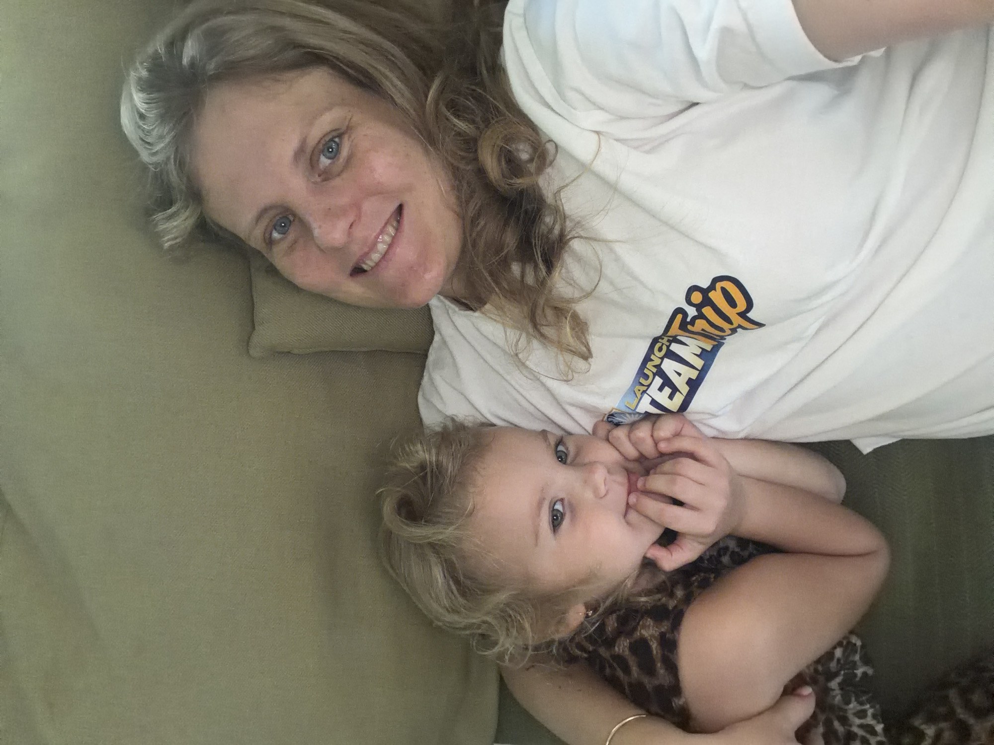 A woman and a young child are lying on a couch, appearing to be taking a selfie. The woman has blonde hair and blue eyes and is smiling at the camera. She is wearing a white t-shirt with a graphic design. The child has blonde hair and green eyes and is leaning into the woman. The child is wearing a leopard print top. They are in a dimly lit room with a tan-colored couch. The image is taken from a high angle, looking down at the two subjects. The background appears to be a wall or another part of the couch.