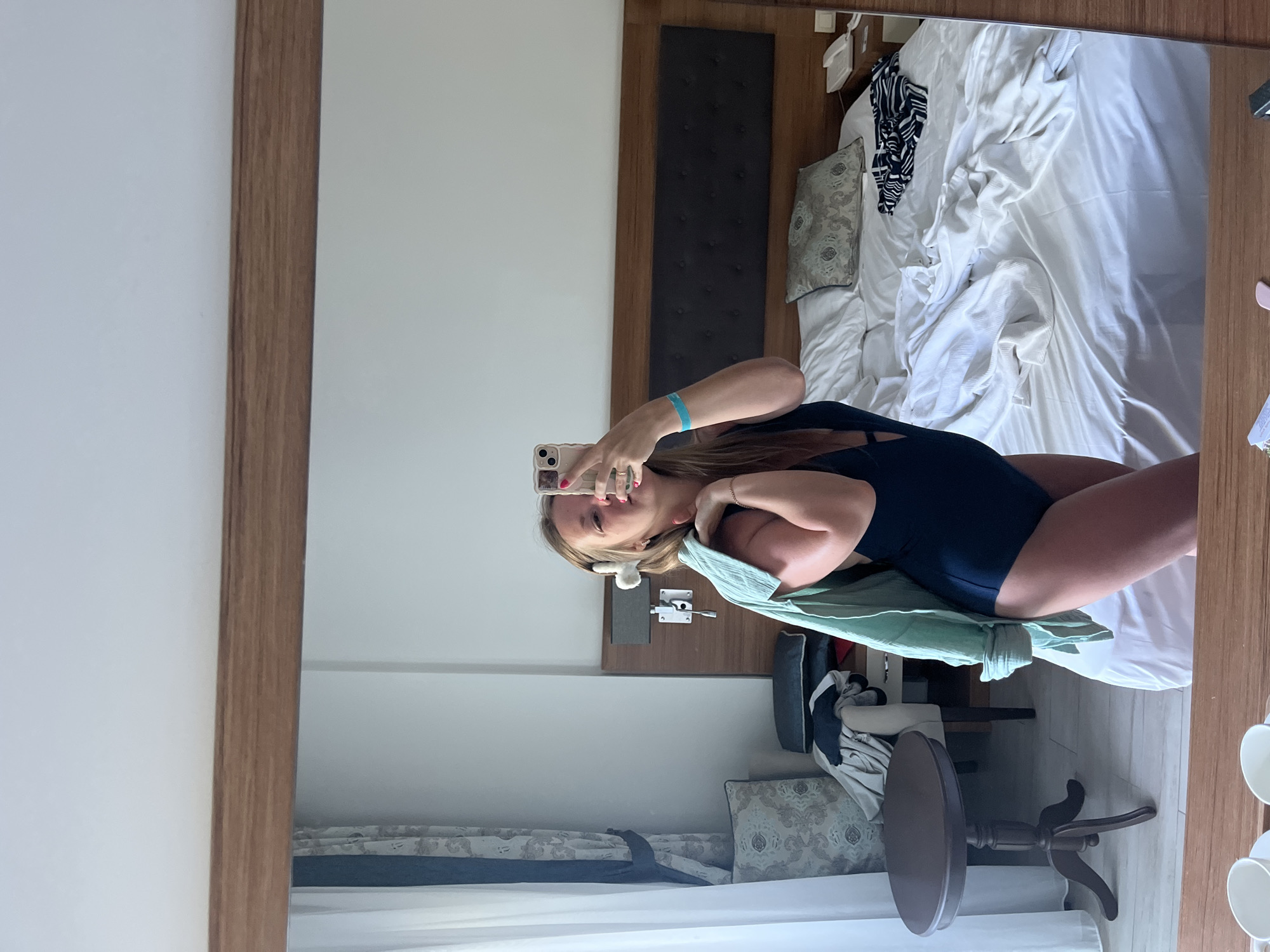 A young Caucasian woman is standing in a hotel room in Alanya, Turkey, taking a mirror selfie. She is wearing a navy blue one-piece swimsuit and has a light blue open shirt draped over her shoulders. Her hair is blonde and tied up with two white fluffy accessories. She is holding a smartphone in her right hand to take the picture. A turquoise bracelet is visible on her left wrist.
The room appears to be a bedroom with a large bed covered in white linens, which looks somewhat disheveled. A dark wooden headboard is behind the bed, with a bedside lamp attached to the wall. To the left of the bed, there is a round, dark wooden side table next to an armchair and a window with white curtains and decorative drapes. Clothes are piled on the armchair and floor. In the reflection of the mirror, a wooden framed shelf with a white object and a dark object is visible on the wall near the headboard. The floor is tiled. In the foreground reflection, two white cups on saucers are visible on a wooden surface, suggesting a desk or dresser. The overall impression is that of a casual, vacation-like setting.