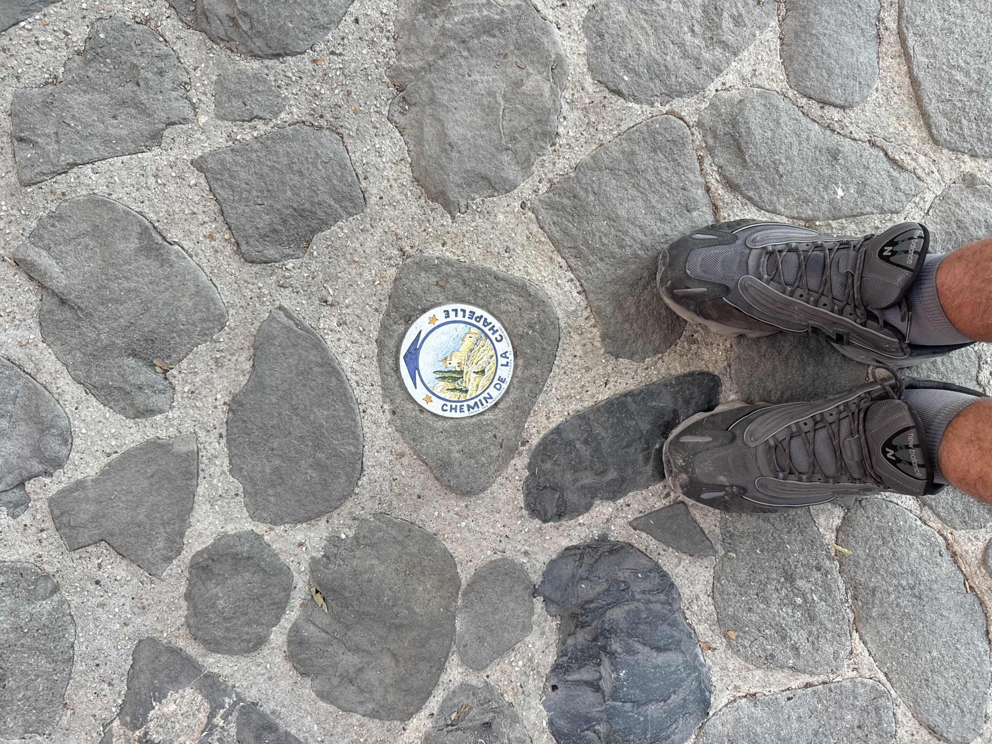 The image shows a close-up view of a cobblestone path in Moustiers-Sainte-Marie, France. A circular sign with an illustration of a village and mountains is embedded in the ground. The sign reads "CHEMIN DE LA CHAPELLE" and "MOUSTIERS S-STE-MARIE". A pair of grey athletic shoes and a portion of a person's legs are visible at the bottom of the frame, indicating the viewer is standing on the path. The stones are irregular in shape and size, and are set in a grey concrete mortar. There are also small pebbles and a few scattered leaves on the ground.