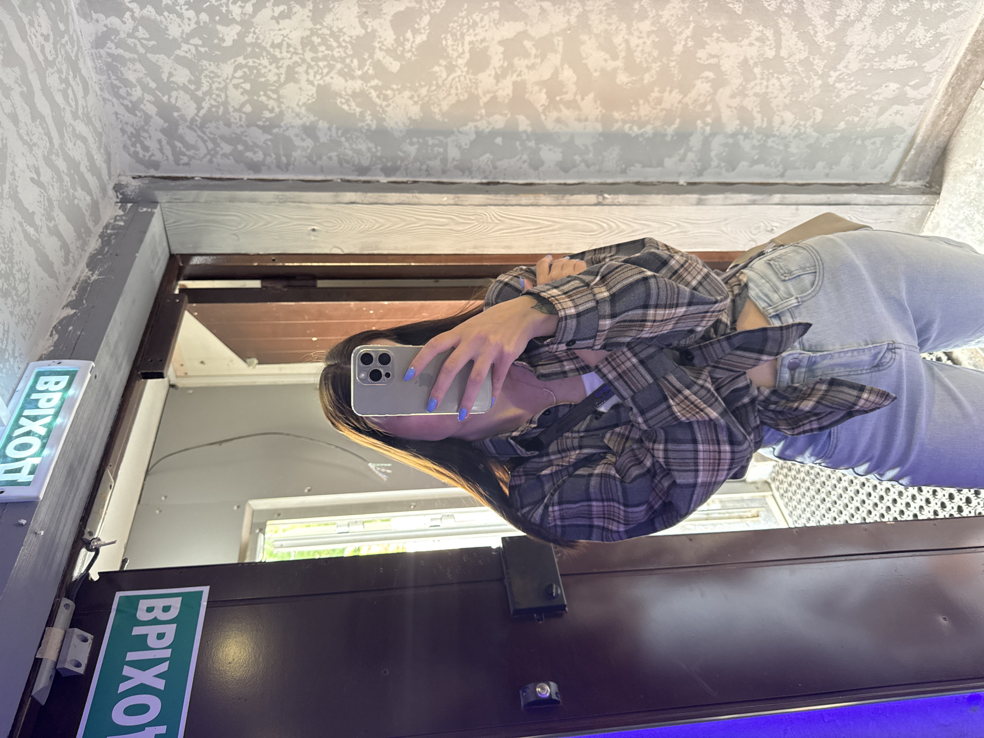 A young woman stands in the doorway of a building in Chaikovsky, Russia, taking a selfie. The building is likely a public space, possibly a business or institution, as indicated by the illuminated "EXIT" sign in Russian on the door frame. The door is dark brown, possibly painted, with visible hardware. The woman is wearing light-colored jeans and a plaid shirt tied at the waist. The ceiling has a textured surface, and the walls are painted with a patterned design. Through the doorway, a glimpse of the outside suggests daylight and greenery.