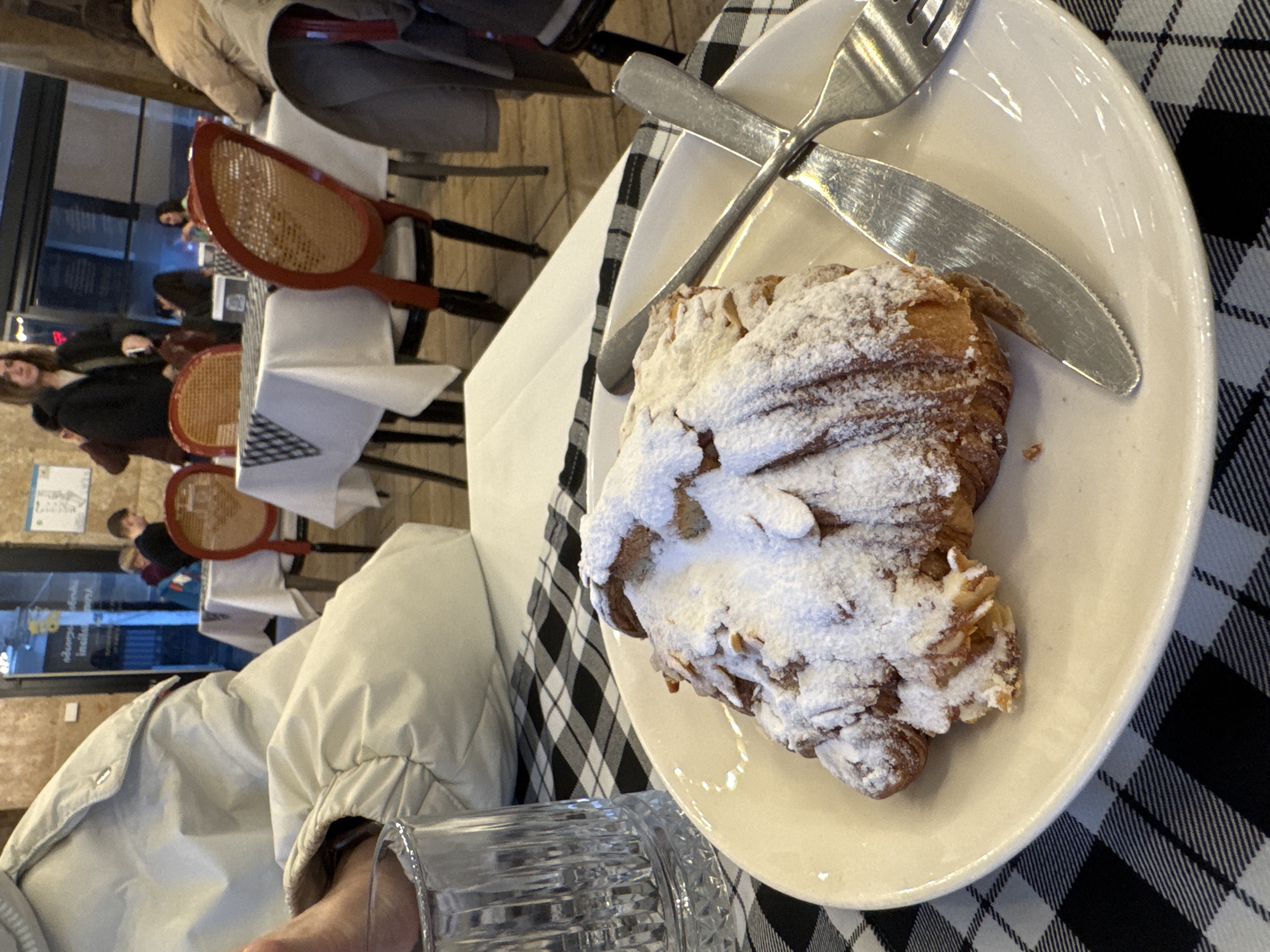The image captures an indoor scene, likely a cafe or restaurant, presented as if rotated 90 degrees counter-clockwise from its upright position. In the foreground, prominently featured on an oval white plate, is a large, golden-brown croissant. It is generously dusted with powdered sugar and topped with slivered almonds. A silver knife with a serrated edge and a matching fork rest across the top of the plate. The plate sits on a black and white checkered tablecloth. To the left, a person's arm, clad in a light-colored puffer-style jacket, is visible, holding a clear, cut-glass tumbler.
In the blurred background, several individuals are present, seated at tables or moving within the space. These tables are covered with white tablecloths and are accompanied by chairs featuring distinctive red frames and woven cane seats. The overall ambiance suggests a casual dining or social setting. The lighting is soft and ambient, indicative of an indoor environment with either artificial lighting or diffused natural light. Some indistinct, blurry text can be seen in reflections on what appears to be a window or glass partition in the far background, but it is not legible. The activity taking place is dining or socializing within the cafe.