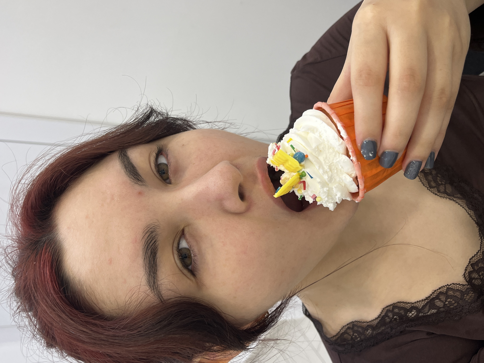 A close-up photograph taken in Neftekamsk, Russia, depicts a female individual in the act of consuming a cupcake. The subject has reddish-brown hair and brown eyes, looking directly at the camera. Her mouth is open, with the top portion of a white-frosted cupcake partially inserted. The cupcake features an orange wrapper with a pink rim, topped with white frosting, colorful sprinkles (red, blue, green, yellow), and a yellow butterfly-shaped decoration. Her right hand holds the cupcake, displaying fingernails painted in a dark grey, glittered polish. She is wearing a dark brown garment with black lace trim along the neckline. Visible on her skin are minor red blemishes on the forehead and cheek. The background consists of an undifferentiated, light-colored wall.