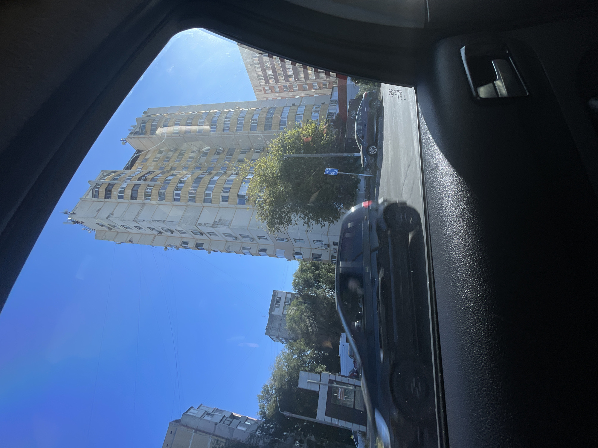 This image captures a street scene in Rostov-on-Don, Russia, likely taken from inside a vehicle. The foreground is dominated by the interior of a car, showing a textured black armrest and a car door handle. Through the window, a tall, pale yellow apartment building with many balconies is visible against a clear blue sky. To the left, another apartment building, more grey in color, stands partially obscured by trees. The street is visible with several cars, including a black hatchback in motion in the mid-ground and a dark sedan parked further down. A traffic light and street signs are also present. The overall impression is a daytime urban environment, with residential buildings and everyday city traffic.