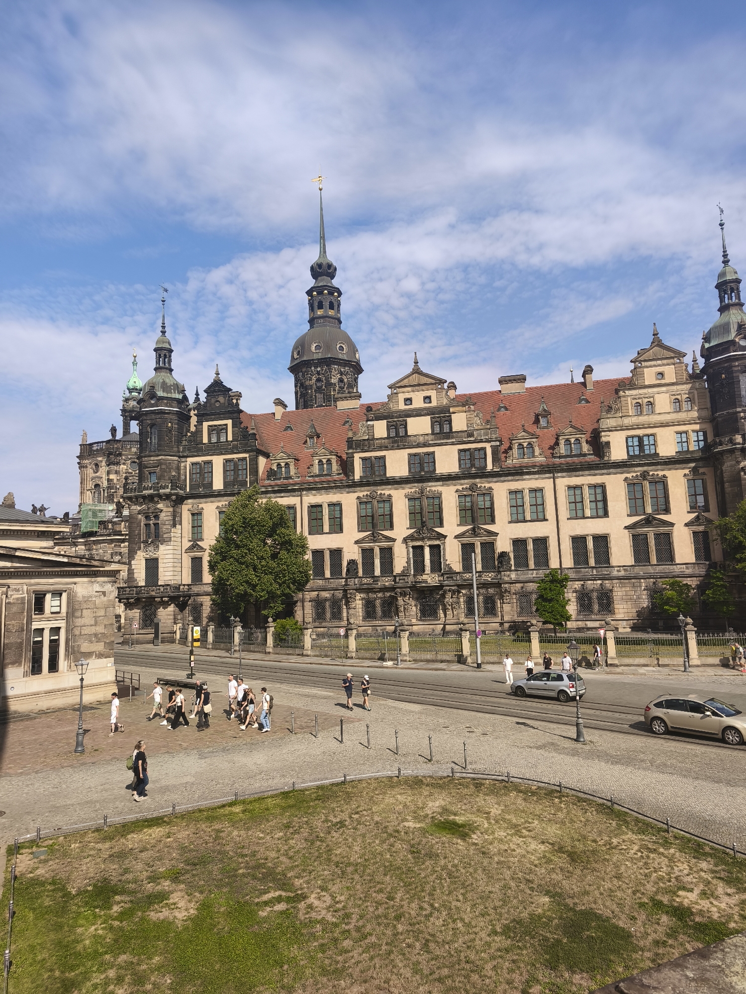 The image shows a daytime view of the historic Frauenkirche in Dresden, Germany. The iconic Lutheran church, with its prominent dome and spires, dominates the background. In the foreground, a cobblestone plaza is bustling with activity. A group of people are walking across the plaza, some in small clusters, while others walk alone. Several cars are visible on the street adjacent to the plaza, and tram tracks are embedded in the road. The sky is a bright blue with scattered white clouds, suggesting fair weather. The architecture of the surrounding buildings is ornate and historic, typical of Dresden's Baroque style. A patch of dry, green grass is visible in the lower right corner of the image.
