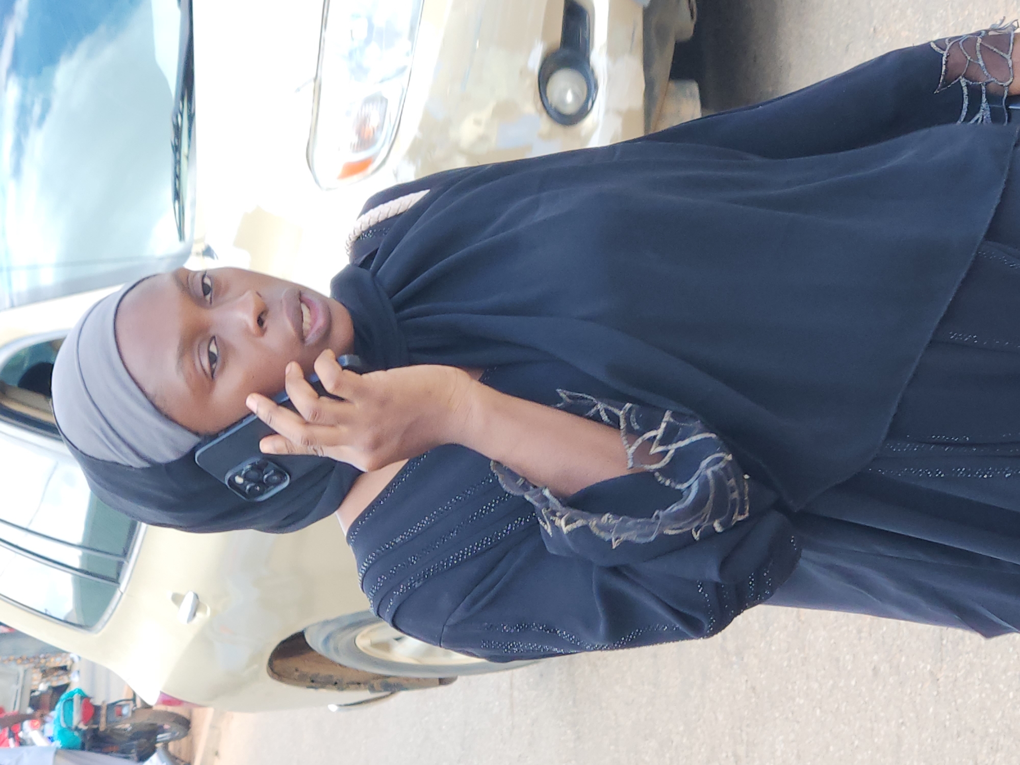 A woman wearing a black abaya and grey hijab is standing on an unpaved road in front of a light-colored SUV. She is holding a black smartphone to her right ear and appears to be speaking. The SUV is partially visible and appears to be dusty. In the background, a motorcycle with a blue bag is also visible, along with other out-of-focus figures and objects, suggesting a busy street scene. The sky is cloudy.