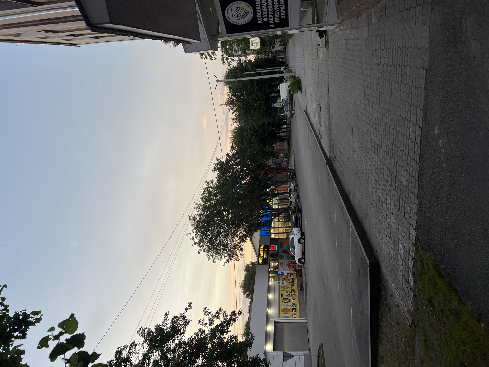 The image shows a street scene in an urban environment, likely during twilight or dusk, as indicated by the soft, diffused light and the pale, muted colors of the sky. On the left, a modern, light-colored building with a white facade and large glass windows appears to be a commercial establishment, possibly a supermarket or a retail store, as suggested by illuminated signs and displays of products. Several cars are parked in a lot in front of this building, including a white SUV and a darker sedan.
To the right, a paved sidewalk with a distinctive interlocking stone pattern runs alongside the street. Adjacent to the sidewalk, another building, taller and with a more traditional architectural style, is partially visible on the far right. This building has a light-colored facade with several windows and a prominent overhang. A sign on this building displays text and a logo, which appears to be related to an association or organization.
The street itself is paved and appears to be relatively calm, with limited traffic. Several trees with dense foliage are scattered throughout the scene, lining the street and bordering the buildings, adding a natural element to the urban landscape. Power lines are visible crisscrossing the sky. The overall impression is one of a quiet street at the end of the day, with shops and businesses still lit and operational.