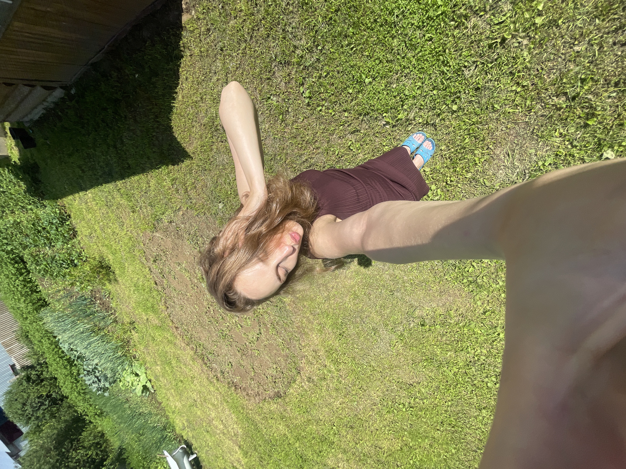 A young woman is captured in a high-angle selfie, lying on her back in a grassy yard with patches of dirt. She is wearing a dark red, ribbed dress and blue sandals. Her eyes are closed, and her hands are placed behind her head, with her hair spread out around her. The background features a garden with rows of green vegetables, lush trees, and a wooden building. The sun casts a strong shadow to the right of the woman. The scene appears to be a private outdoor space, possibly a backyard in the village of Sharkan, Russia, given the presence of a garden and a wooden structure suggestive of a dacha or rural home.