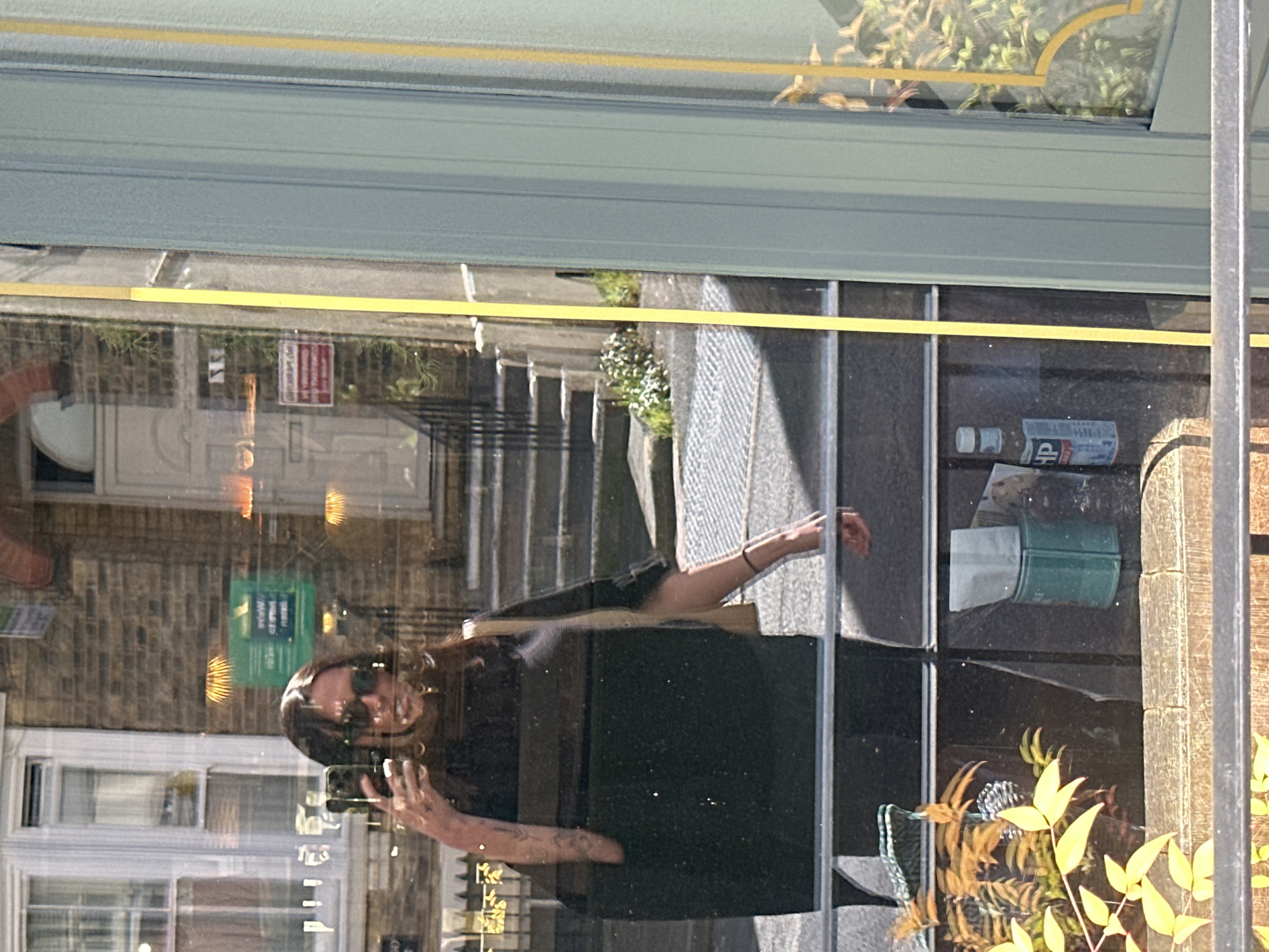 The image is a reflection in a shop window, showing a woman taking a photo of herself. The reflection shows the exterior of a building across the street, the woman's reflection, and items inside the shop window, including some newspapers, a can of soup, plants, and other miscellaneous objects. The reflection of the building suggests a brick façade with white-framed windows. Based on the reflections in the shop window, and the architecture of the building visible in the reflection, it is probable that the location is within Tunbridge Wells, United Kingdom.