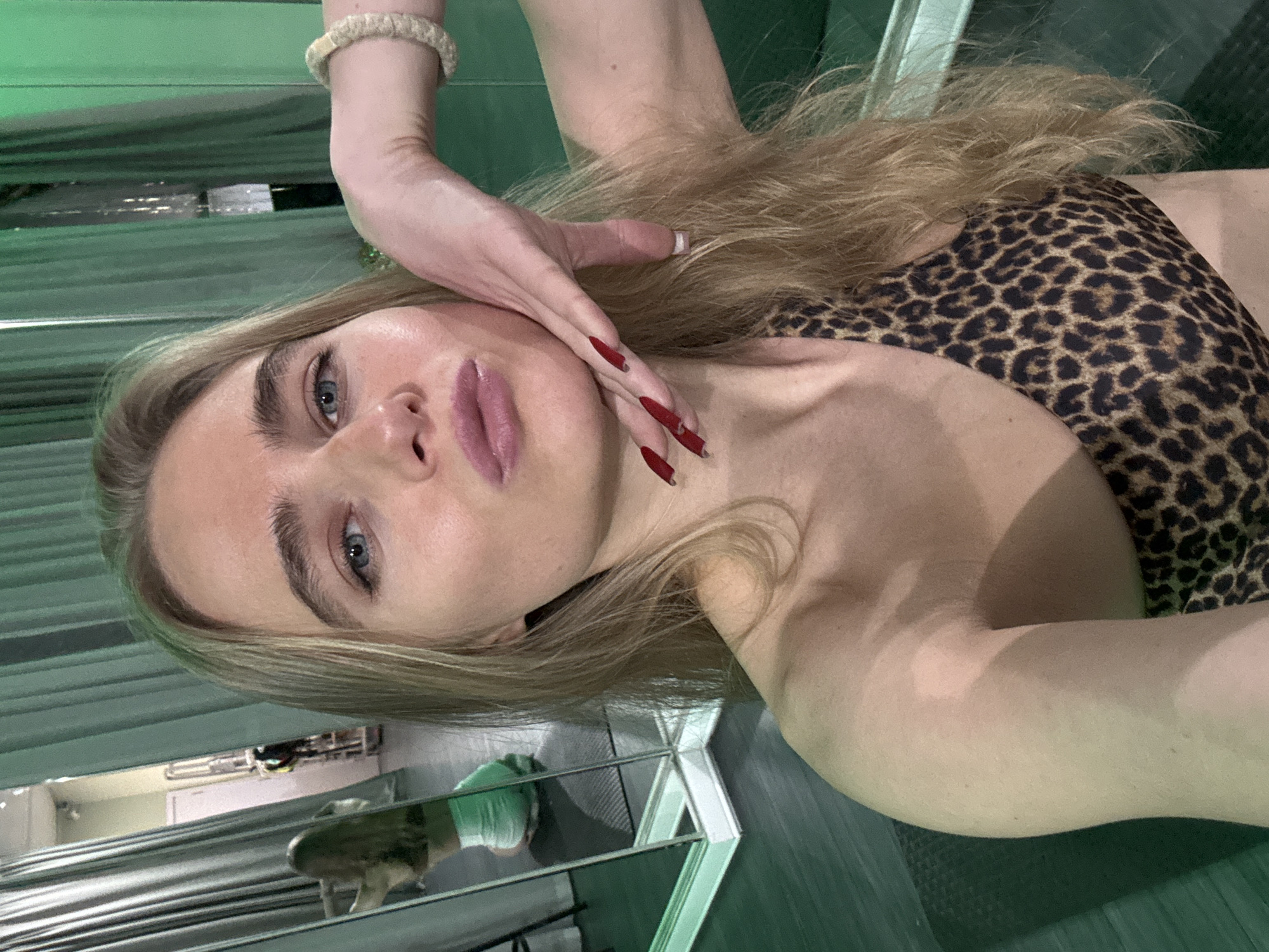 A woman is posing indoors, wearing a leopard-print top. One hand is touching her face and the other is raised with a bracelet. Her hair is blonde and long. The room appears to have reflective surfaces.