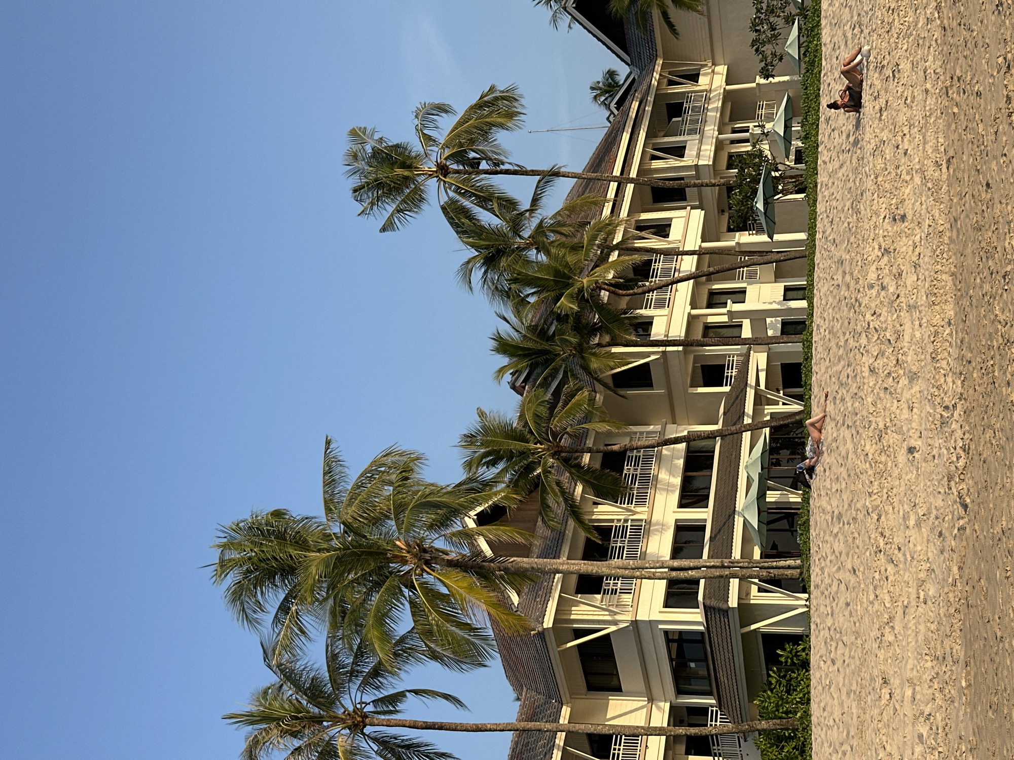A panoramic image captures a beachfront scene in Choeng Thale, Thailand, under a clear blue sky. The foreground consists of an expansive area of light-colored sand. Two individuals are present on the sand: one person is lying supine on the left, wearing dark clothing; a second person is lying prone on their stomach, propped on elbows, wearing dark attire with visible white shoes, positioned towards the right. Behind the sand, a multi-story, cream-colored building with dark tiled roofs and multiple balconies extends across the midground. Numerous tall palm trees are interspersed throughout the scene, several standing directly in front of the building. Multiple light-colored beach umbrellas are visible on a manicured green lawn situated between the sand and the building. The scene is illuminated by natural light, casting subtle shadows.