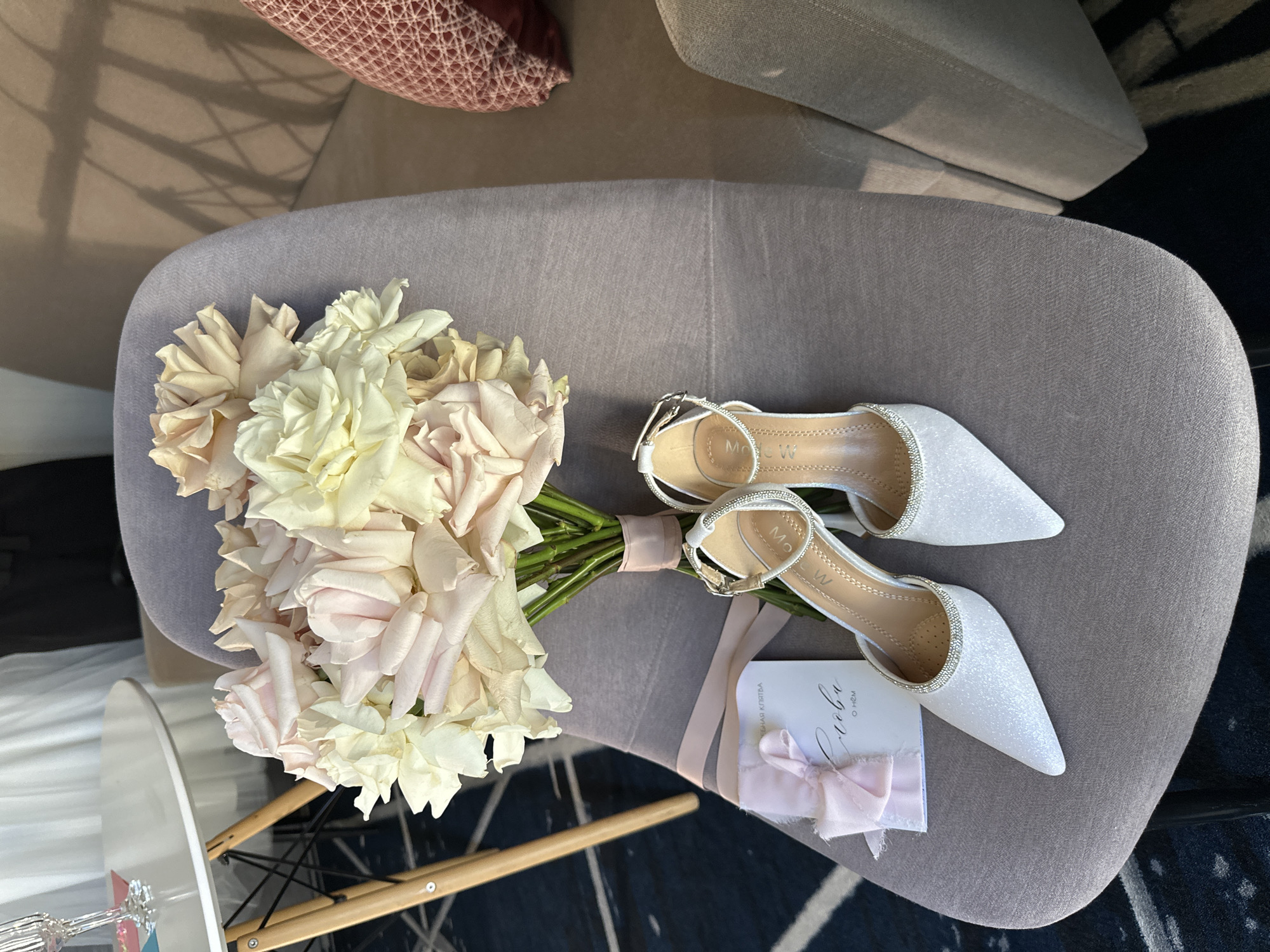 A close-up, eye-level, horizontal, outdoor shot in Novosibirsk, Russia, depicts a pair of white, pointed-toe, glittery high-heeled sandals with ankle straps and delicate rhinestone embellishments, placed on the seat of a light gray upholstered armchair. To the left of the sandals lies a small, white card tied with a pale pink ribbon, on which the word "Слова" (meaning "Words") is written in elegant calligraphy, suggesting it is part of a wedding or special occasion setup. Behind the card and sandals, a bouquet of roses in varying shades of cream, pale pink, and light yellow is arranged, tied with a matching pale pink ribbon. The bouquet’s stems are visible, and the roses appear fresh and full. The armchair is situated on a dark patterned rug. In the background, a white circular table with a mid-century modern wooden base is partially visible, with a clear glass on its surface. To the far right, the armrest and back of another upholstered chair in a taupe color are seen, along with a textured, pink throw pillow. The overall scene evokes a sense of a celebratory event, likely a wedding, with elements prepared for a bride.