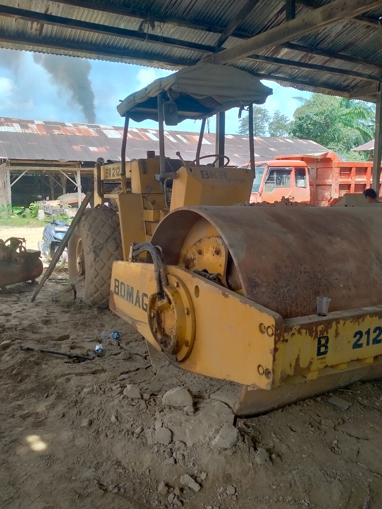 The image depicts a yellow Bomag BW 212 road roller parked on a dirt surface under a corrugated metal roof in Menanggini, Indonesia. A dump truck is visible behind the road roller. Other equipment and a motorcycle are in the background, partially obscured. Smoke is visible in the distance.