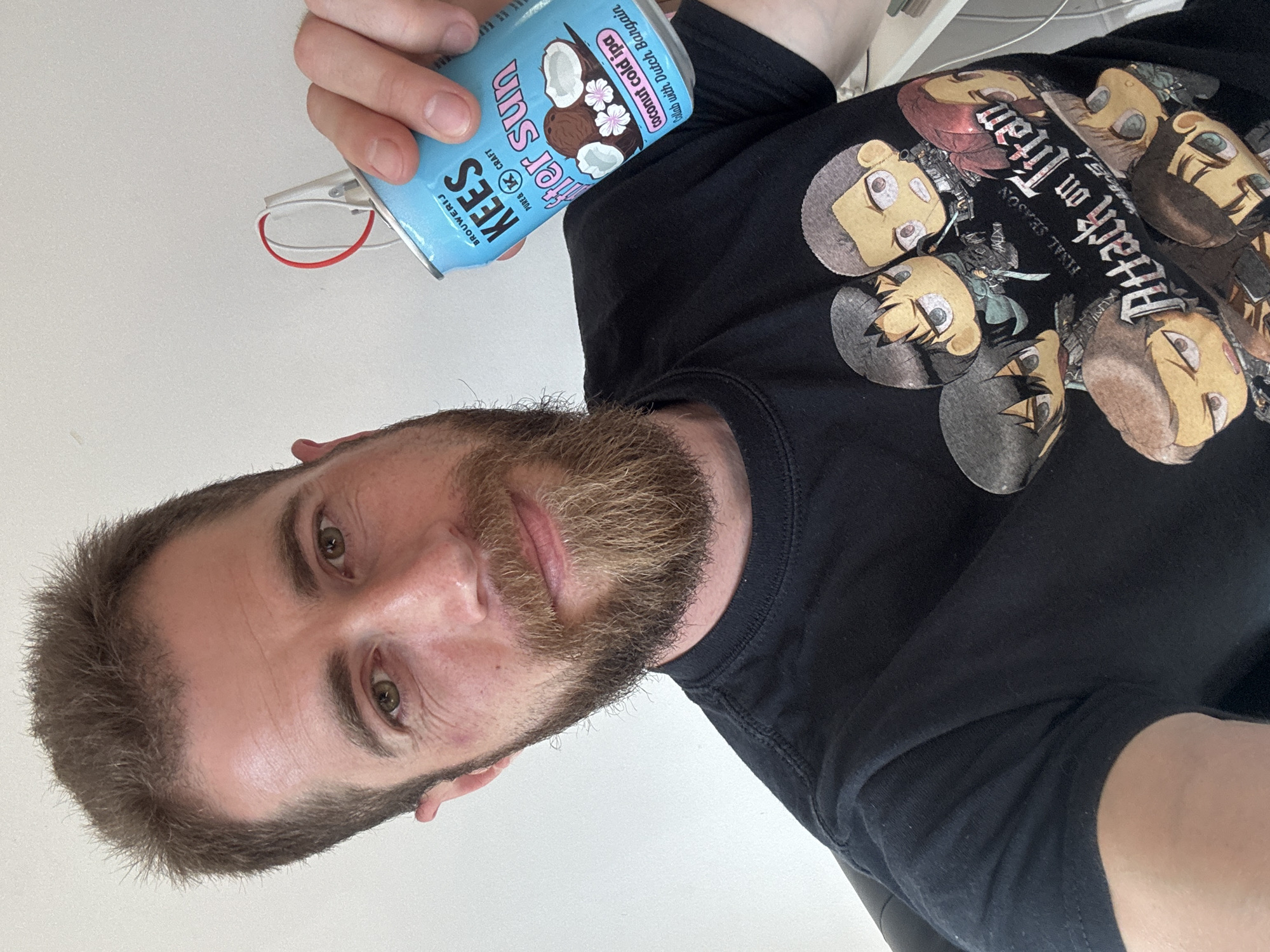 A man is holding a can of KEES After Sun coconut cold IPA in his left hand. He is wearing a black t-shirt with "Attack on Titan" printed on it. He has short brown hair, a beard, and green eyes. The can reads "Brouwerij KEES" and describes itself as a "pure & K craft" beer. The can also notes "Collab with Dutch Bargain." The background is out of focus and provides no clues to the location of Budva, Montenegro.