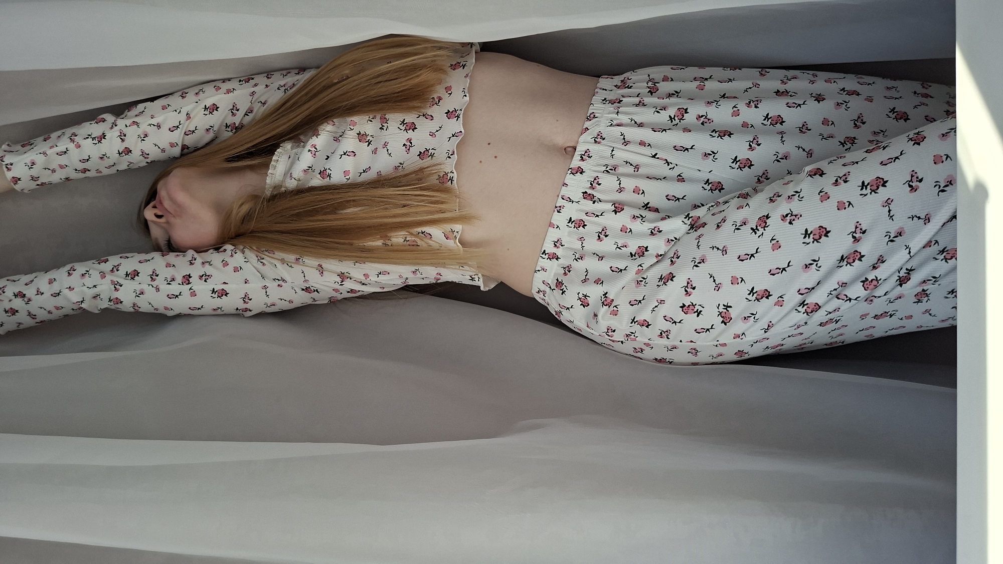 An overhead shot captures a person, likely female, with long, flowing blonde hair, lying supine in a serene pose. They are dressed in a two-piece white pajama set adorned with a repeating pattern of small pink and black roses. The long-sleeved crop top reveals a portion of their midriff, which features a few small moles. The matching pajama pants cover their legs.
The person's arms are stretched out upwards and slightly to the sides, parallel to their body. Their head is turned slightly to the right, with their face partially obscured by their hair and the angle, suggesting a relaxed state.
They are situated within a soft, cocoon-like enclosure formed by two layers of light-colored, semi-transparent fabric, possibly white or light grey. The lower fabric appears to be a flat surface, while the upper fabric drapes gently around and over the subject, creating soft folds. A darker, flat surface is visible beneath the person, contrasting with the lighter fabrics. Soft, natural light originates from the right side of the frame, casting subtle shadows and highlighting the texture of the fabrics. The overall impression is one of tranquility and comfort. No visible text is present in the image. This scene is set in Chaikovsky, Russia.