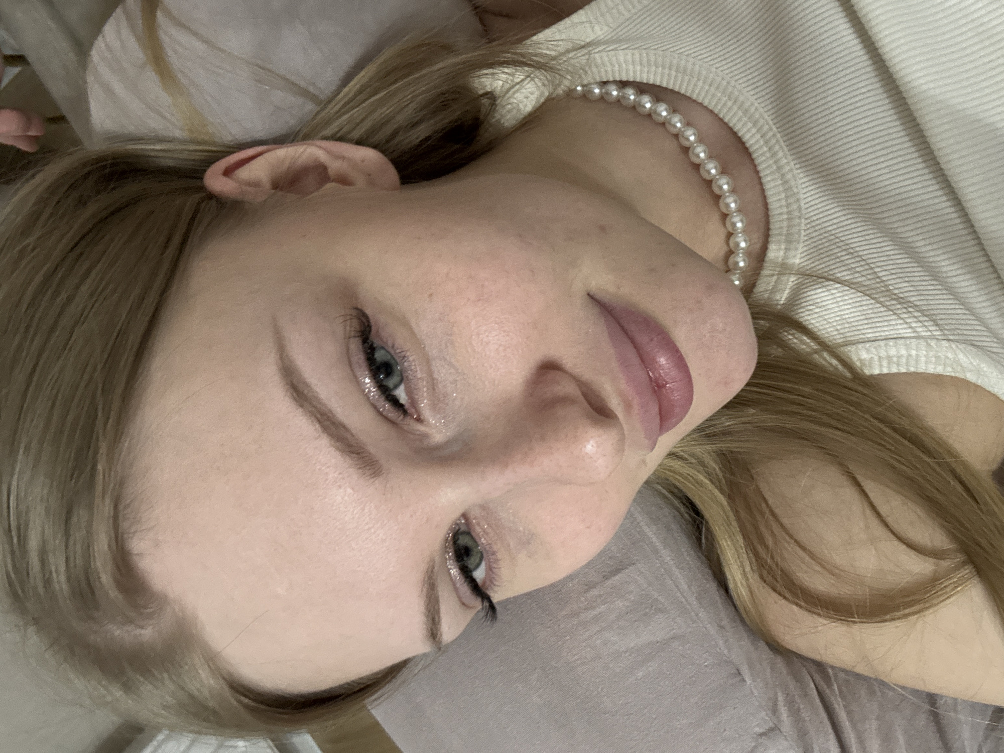 A close-up portrait features a young woman lying down, her head tilted to the right. Her light blonde hair, parted on her left, frames her face and flows onto a light grey surface, likely a pillow or bedding. She has fair skin and expressive blue-green eyes, accentuated with dark eyeliner, mascara, and a shimmering, glittery eyeshadow. Her lips are a soft, glossy pink.
Her gaze is directed slightly upwards and to the viewer's left, creating a calm and serene expression. She wears a classic pearl necklace, visible around her neck, peeking out from a white, ribbed top. The overall setting appears to be indoors, suggesting a relaxed environment like a bedroom. No text is visible in the image.