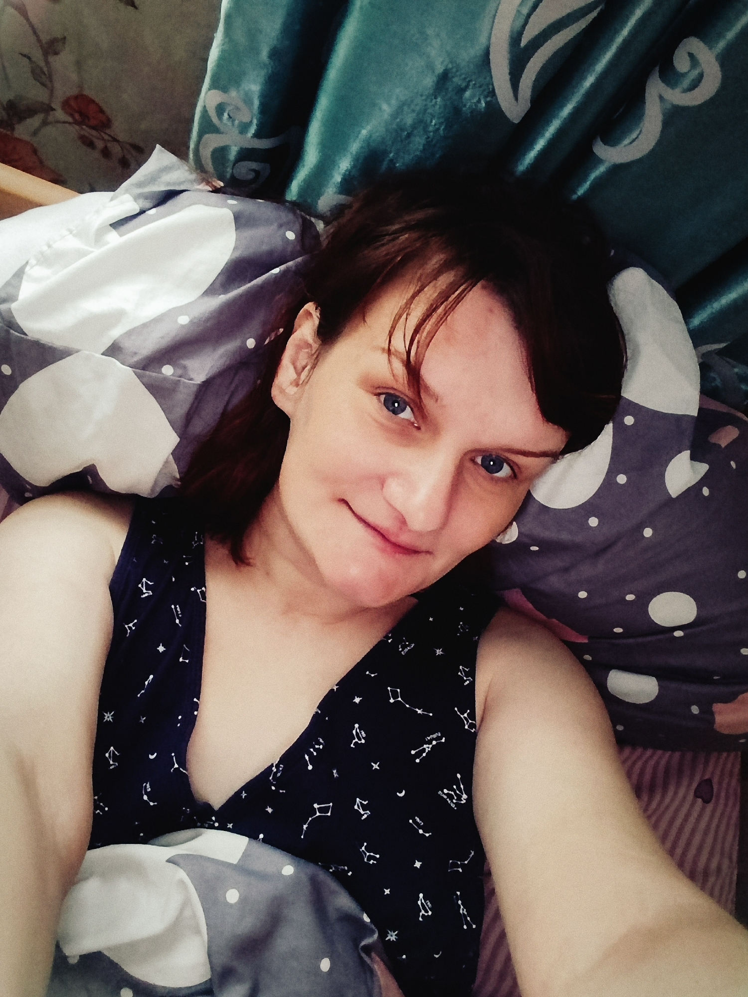 A person is lying down, presumably in a bed, looking at the camera. The person has fair skin, blue eyes, and short brown hair with bangs. They are wearing a sleeveless top with a dark background and a constellation pattern. The person's head rests on a patterned pillow of gray and white. A teal curtain is visible behind the pillow. The location cannot be determined from the image content.
