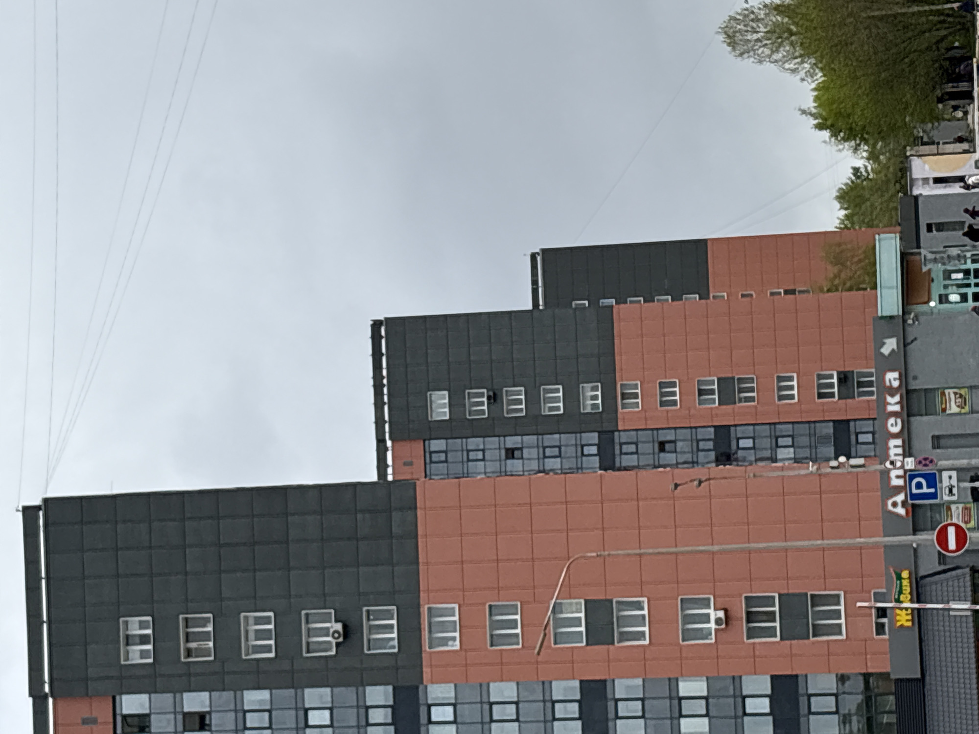 The image depicts a multi-story building in Perm, Russia. The building has a stepped design with alternating sections of gray and reddish-brown paneling. Numerous windows are visible. A sign with the word "Аптека" (pharmacy) is present. Utility wires are strung overhead against a cloudy sky. Trees are visible in the background. Street signs and what appears to be a parking sign are also present. The image is rotated clockwise.