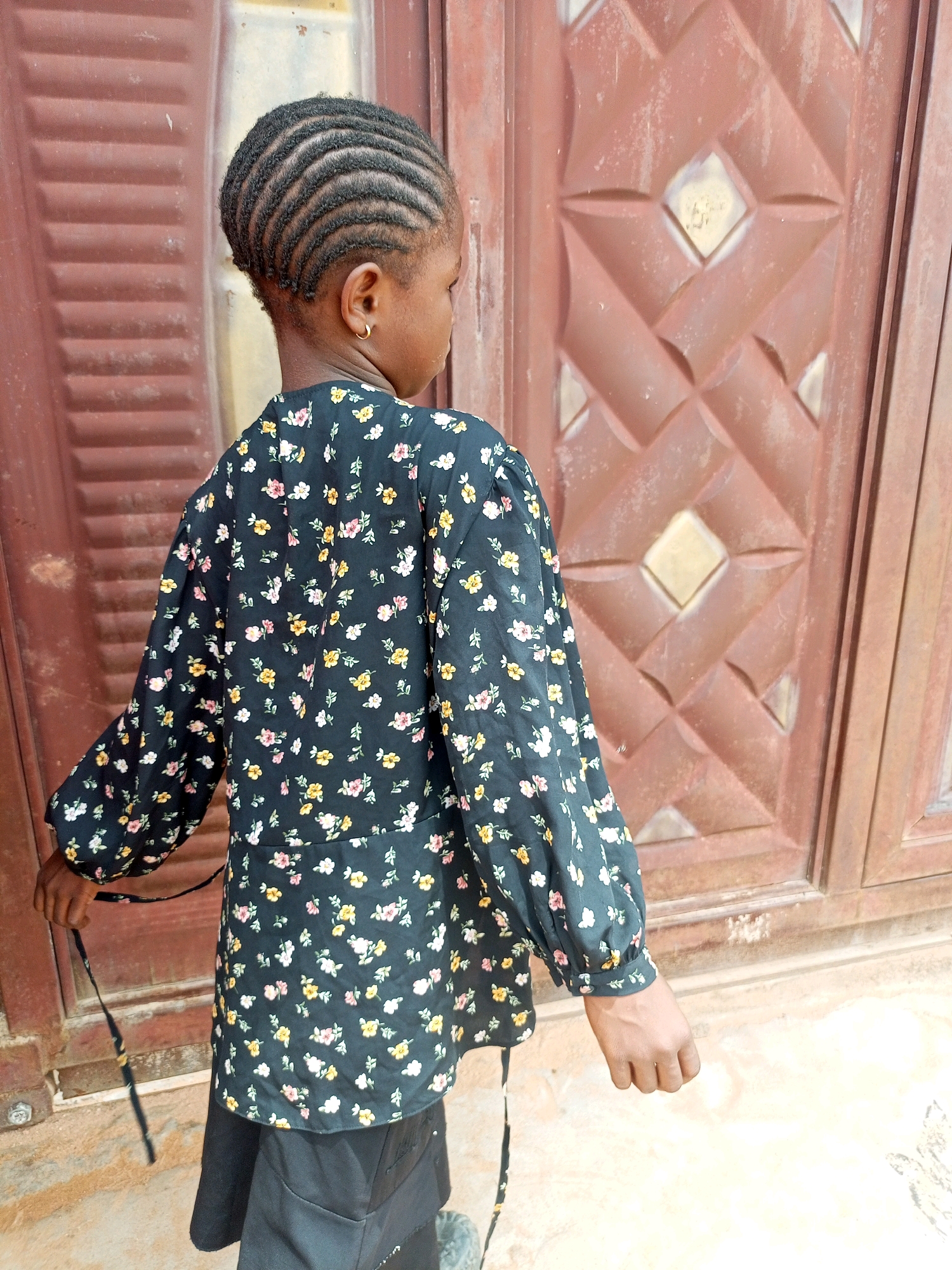 A young Black girl, with braided hair and wearing a floral print blouse and dark pants, stands facing away from the camera in front of a brown, patterned metal door. The door has a ribbed texture and a diamond-shaped pattern. The girl is holding a thin, dark strap in her right hand. The ground in front of the door appears to be concrete or pavement, with some dirt and debris. The overall scene suggests an outdoor setting, possibly in a residential area. The location is identified as Tempure, Nigeria.