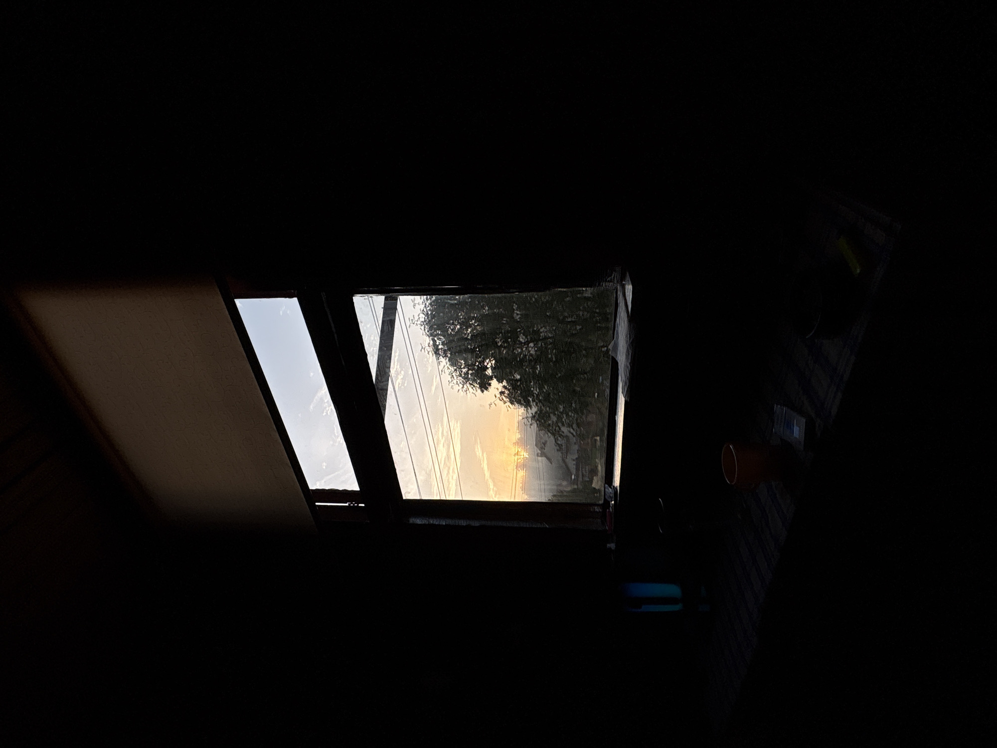 The image depicts a window looking out onto a misty sunrise over a landscape of trees and buildings, possibly in Jāņupe, Latvia. The room is dark, with light streaming in from the window, casting shadows on the floor and a surface below the window. On this surface, a blue cylindrical object, an orange cup, and a pack of cigarettes are visible. The window frame is dark wood, and a light-colored blind is pulled down at the top of the window. The sky is filled with soft, warm light, and the mist obscures the details of the landscape, creating an atmospheric and serene scene.