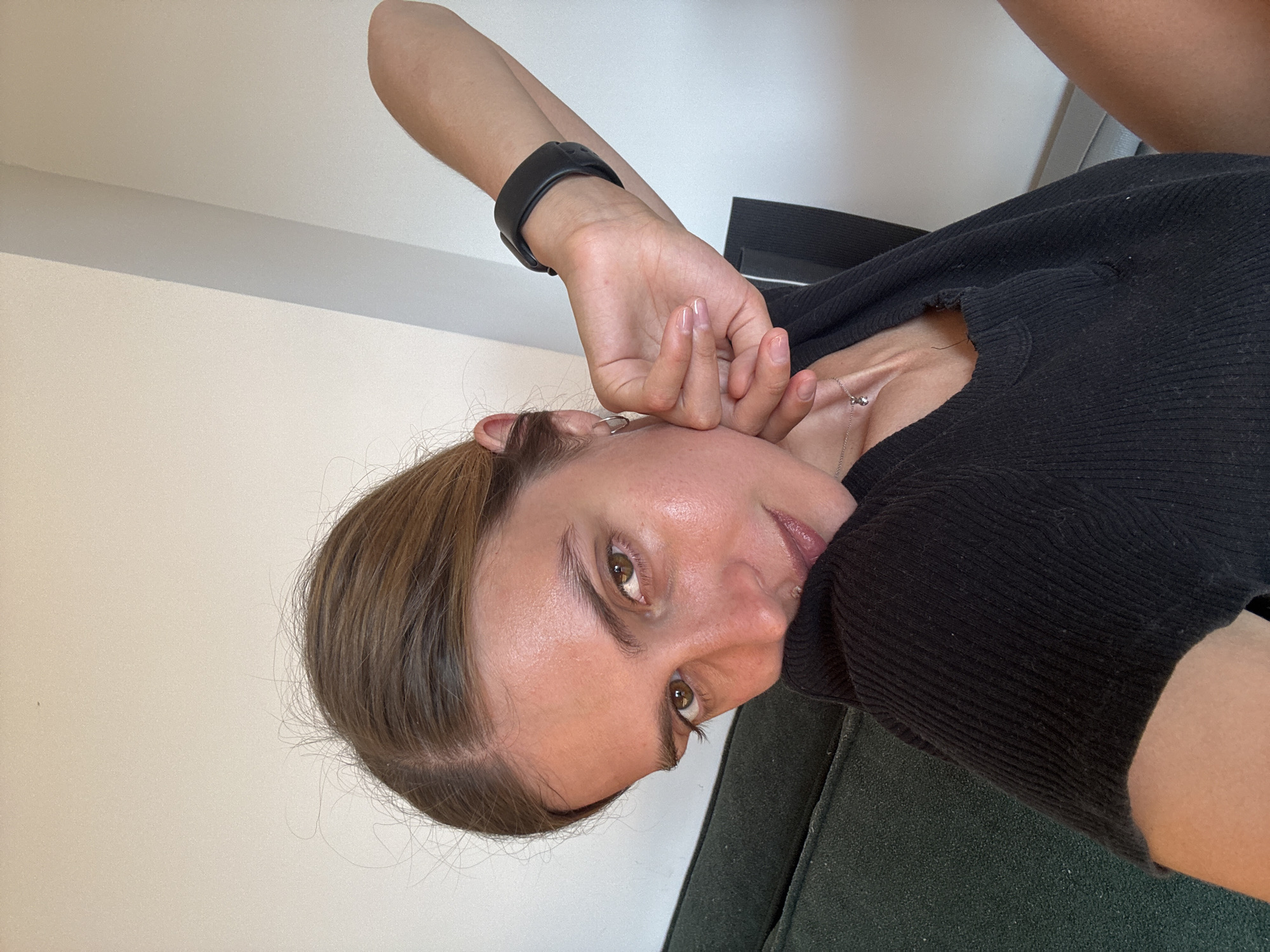The image displays a close-up portrait of a young adult female, captured as if taking a selfie or from an overhead angle, although the picture is rotated 90 degrees clockwise. She has brown hair, fair skin, and brown eyes, with a direct and neutral gaze toward the camera.
She is wearing a black, ribbed long-sleeved top, and a delicate silver chain necklace with a small pendant is visible around her neck. On her left wrist, she sports a black digital watch. Her left hand is raised, with her fingers gently resting near her forehead or temple, partially covering her face. Her nails appear to be manicured with a light, possibly clear or nude, polish.
The setting appears to be indoors, with a plain light-colored wall forming the upper background. A dark green upholstered surface, likely a couch or chair, is visible in the lower part of the frame. The lighting is bright and diffused, characteristic of an indoor daytime scene. There is no visible text in the image.