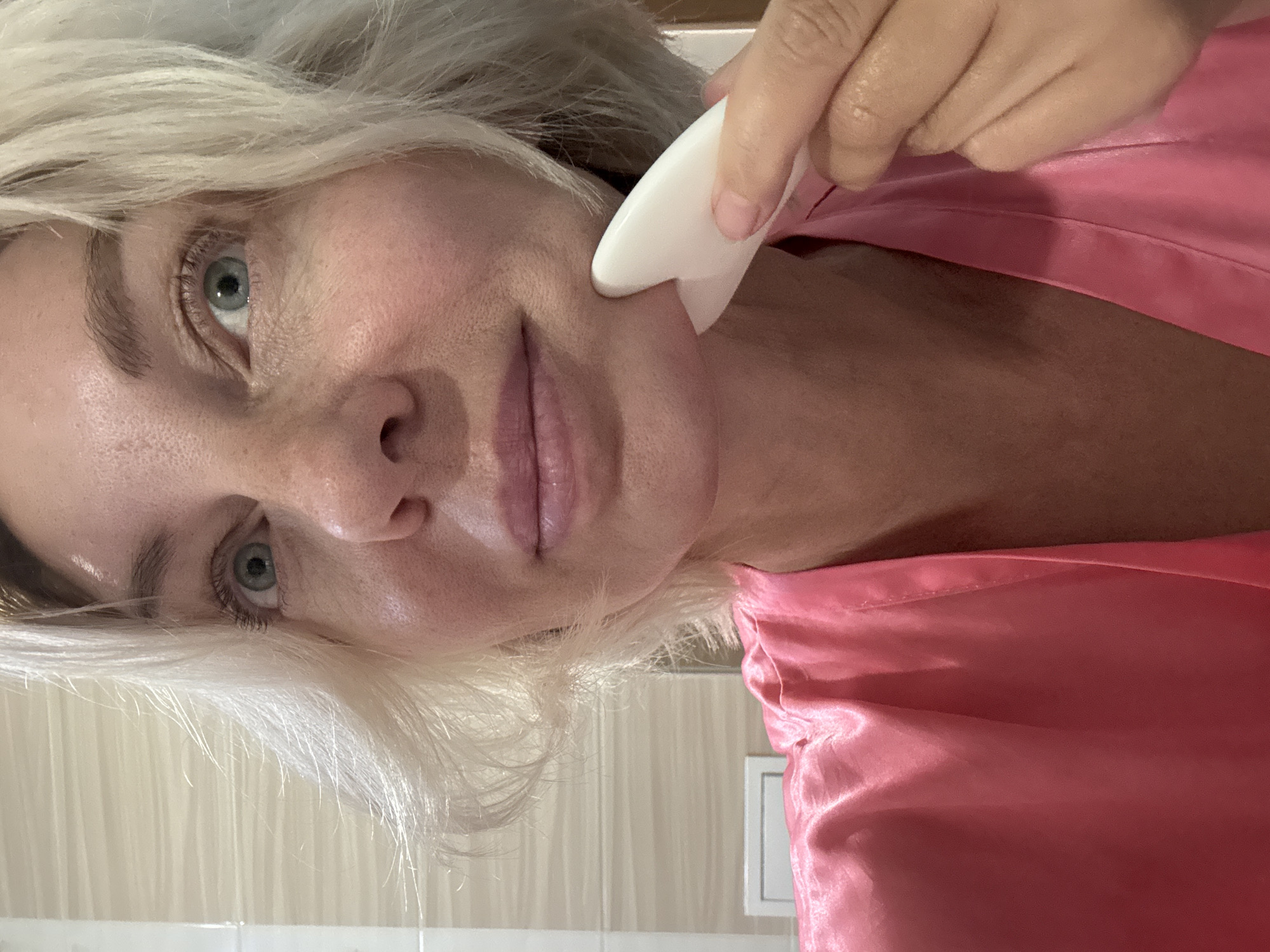 A middle-aged Caucasian woman with blonde hair and light blue eyes is in a bathroom, performing a facial massage with a white gua sha tool on her jawline. She is wearing a pink satin robe. The bathroom appears to be clean and modern, with tiled walls and a white vanity visible in the background. The lighting is soft and natural. The image was captured in a bathroom in Sochi, Russia.