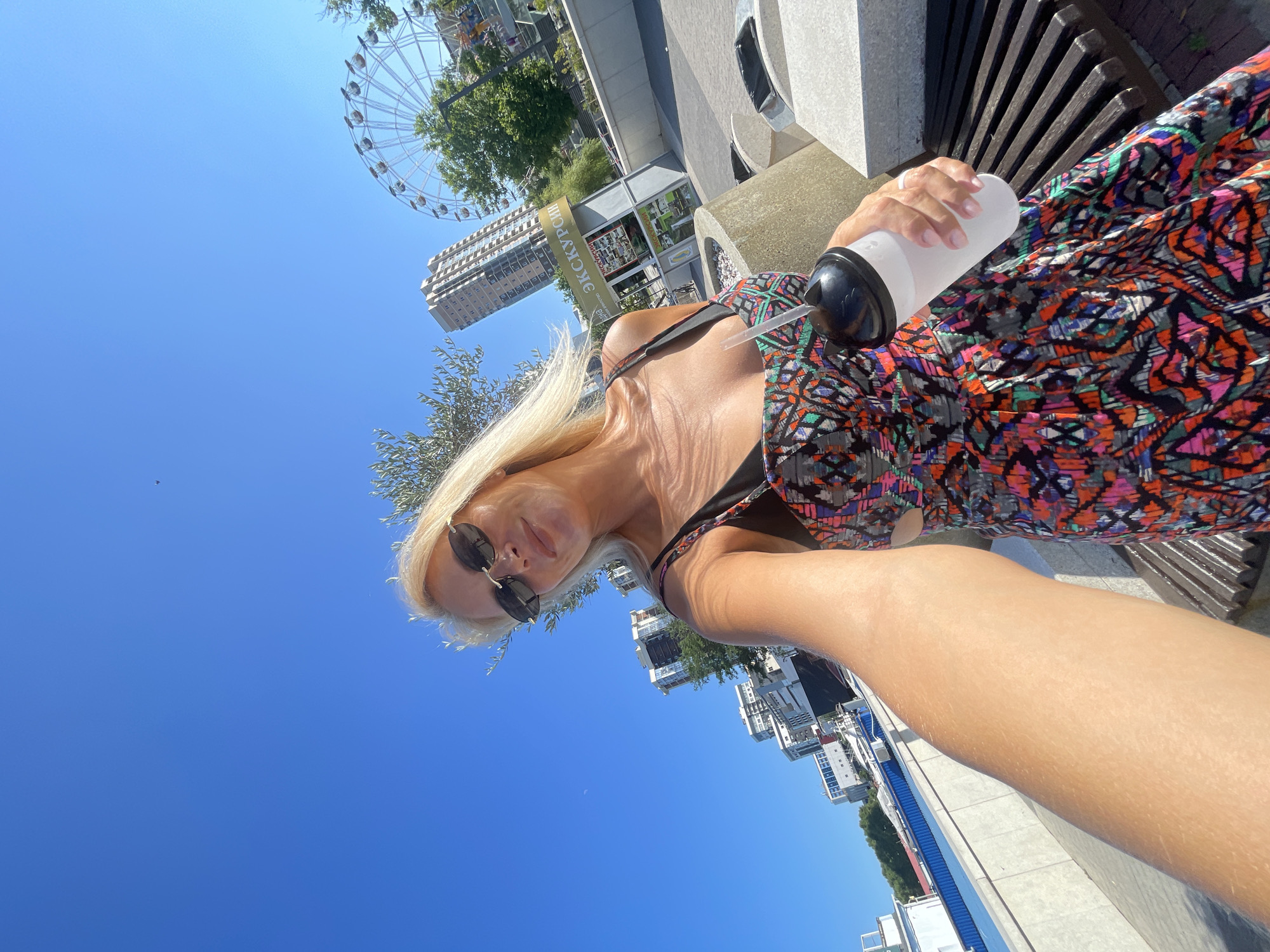 A woman with blonde hair and sunglasses is taking a selfie outdoors, holding a white cup with a straw. She is wearing a colorful, patterned dress and standing on a paved area next to a wooden bench. In the background, there are several tall buildings, a Ferris wheel, trees, and a blue sky. The location appears to be a city waterfront. The text on a kiosk in the background is partially visible and includes the word "ЭКСКУРСИЯ," which means "excursion" in Russian. This suggests the location might be a tourist area.