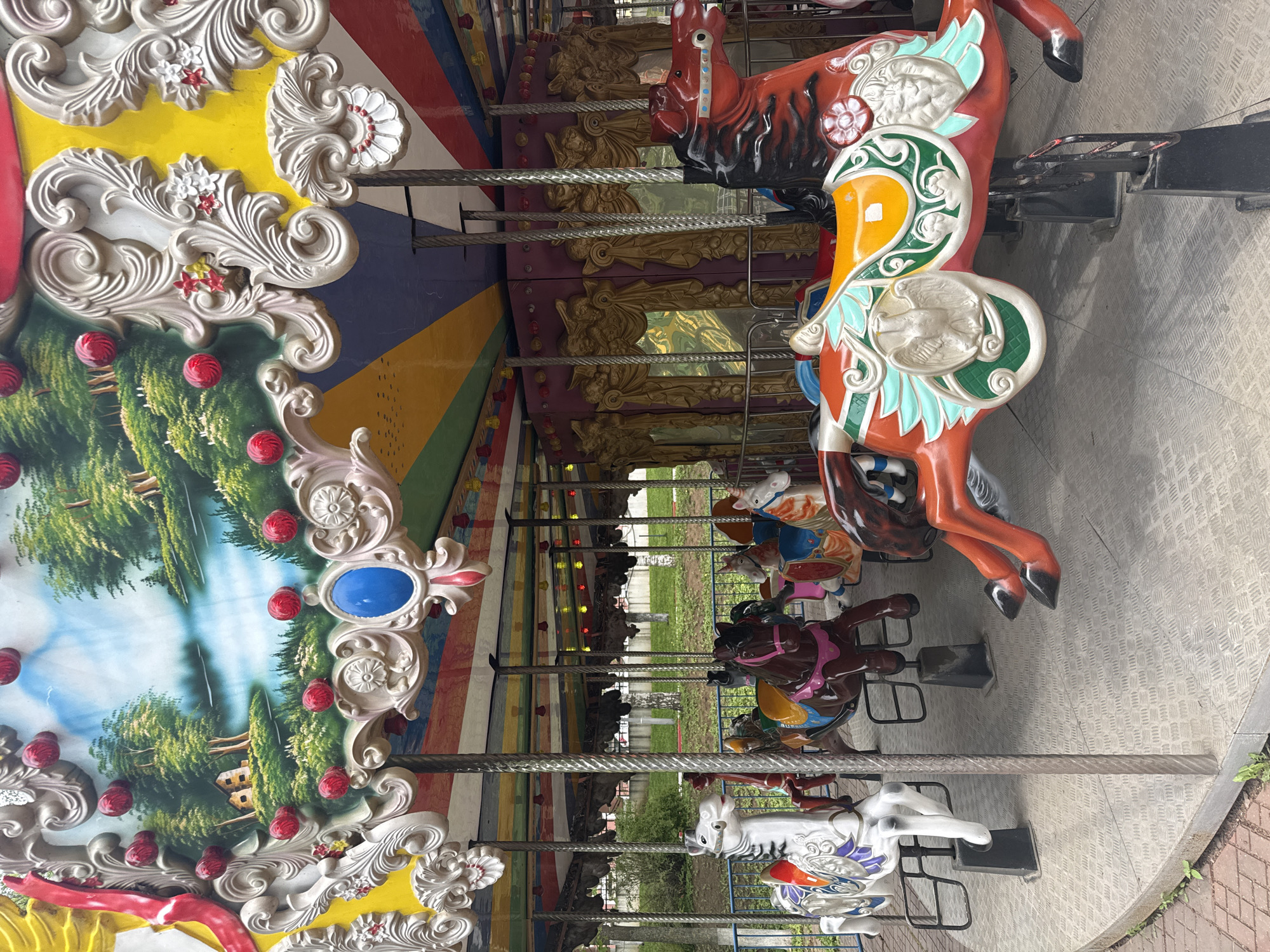 The image depicts a carousel in Izhevsk, Russia. The carousel features ornate decorations, including painted panels with landscape scenes, decorative trim with faux flowers and berries, and colorful striped sections. Rows of carousel horses are visible, each with unique paint schemes and designs. The carousel appears to be situated outdoors, with trees and park-like scenery visible in the background. The ground around the carousel is paved.