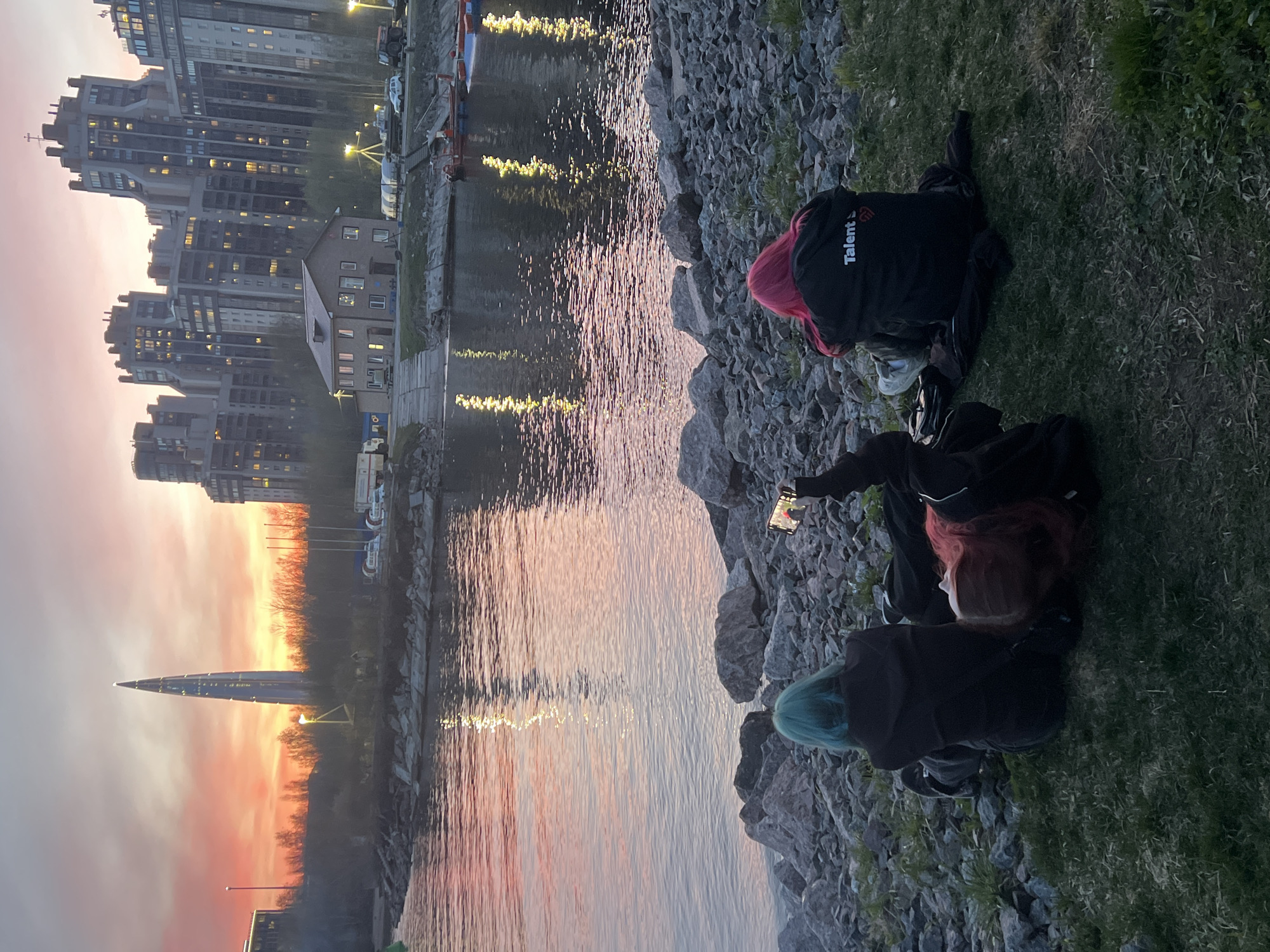 The image captures a scenic waterfront view in Saint Petersburg, Russia, during what appears to be dusk or dawn. The sky is awash with vibrant orange and pink hues, reflecting beautifully on the calm surface of the water.
In the foreground, two individuals are seated on a grassy bank next to a rocky shoreline. One person, positioned to the right, has striking pink hair and is wearing a black jacket with "Talent S" (or similar, partially visible) printed in white on the back. The second person, slightly to the left, has bright teal hair, wears dark clothing, and is holding a smartphone, seemingly capturing the picturesque scene or the sunset/sunrise.
The background features a collection of modern, multi-story buildings lining the far bank of the water, many with illuminated windows. Most notably, the distinctive, tall, and slender Lakhta Center skyscraper is prominently visible on the distant horizon against the colorful sky, serving as a key location cue. Reflections of building lights shimmer on the water's surface. The overall atmosphere appears peaceful and contemplative, with the people enjoying the serene view and warm light.
