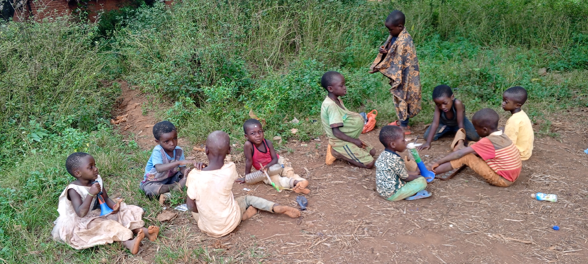 The scene in Ngalula, Democratic Republic of the Congo, features a group of nine children in an outdoor setting. They are positioned on a dirt ground, partially covered with sparse vegetation. To the left of the frame is dense green foliage.
The children are barefoot and wear assorted clothing, including dresses, shirts, and trousers, which appear worn. Several children are seated on the ground, while one child stands wearing a patterned garment. Some children hold blue objects, possibly toys, while others have plastic bottles nearby.
The lighting indicates it's daytime with natural sunlight. The environment appears impoverished, with sparse personal belongings around the children. There is no visible infrastructure or indication of urban development.