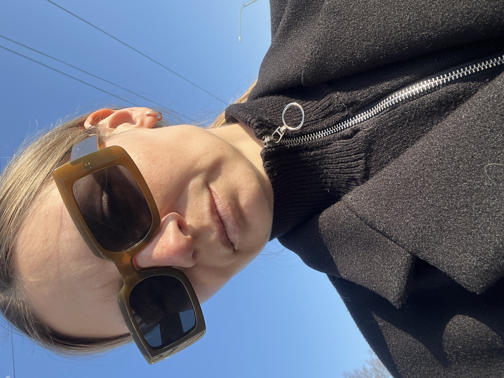 An outdoor, low-angle close-up image depicts an individual, likely female, in the city of Chaikovsky, Russia. The subject, with light-colored hair pulled back, occupies the majority of the frame, facing slightly towards the camera. She wears large, rectangular, olive-green sunglasses with dark lenses, obscuring her eyes. Her nose exhibits freckles. A small, gold-colored hoop earring with visible embellishments is present on her right earlobe. Her lips are closed, presenting a neutral expression. She is dressed in a black, high-collared knitted garment featuring a silver zipper with a circular pull ring, worn beneath a black, textured overcoat with lapels. The background consists primarily of a clear, bright blue sky. Three parallel utility lines are visible in the upper right quadrant. A street light fixture is partially visible on the right side. Faint outlines of bare tree branches are discernible in the lower-left corner.