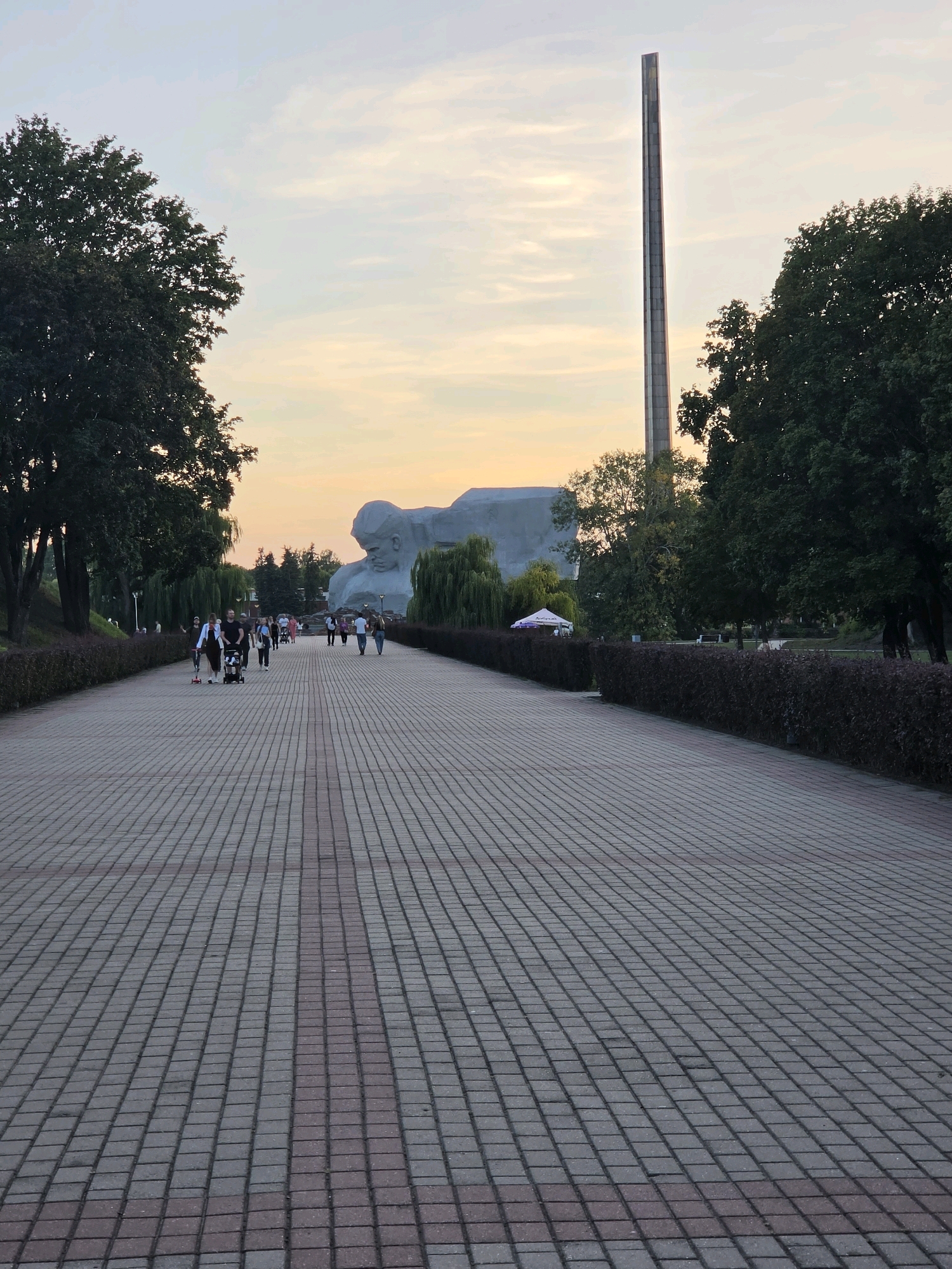 The image depicts a view down a wide, paved pedestrian walkway in Brest, Belarus. The walkway is constructed of interlocking grey and red pavers, creating a striped pattern. On either side of the walkway are neatly trimmed dark hedges. Beyond the hedges, on the left, is a grassy slope with trees and an overhead pedestrian walkway. On the right, beyond the hedges, are more trees and a body of water.
In the background, the sky is a soft gradient of pale orange and yellow, indicative of either sunrise or sunset. Dominating the background is a large, grey, sculptural monument depicting a reclining figure. To the right of the monument, a tall, slender obelisk rises into the sky. Several people are visible walking along the paved walkway in the mid-ground, some with strollers, and others on scooters. In the distance, a small white tent with branding is visible near the water's edge. The overall scene suggests a public park or memorial area, likely the Brest Fortress Memorial Complex, a prominent landmark in Brest, Belarus.