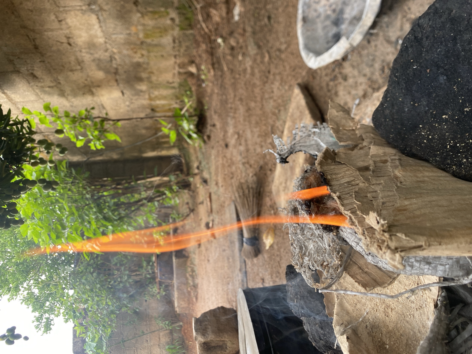 The image depicts an outdoor scene in Bukuru, Nigeria. A fire burns, composed of cardboard, metal, and wood. Near the fire, there is a broom, a black rock, and a metal dish. The surrounding environment includes lush green trees, a wall, and dirt ground. The camera has captured light trails that are orange in color.