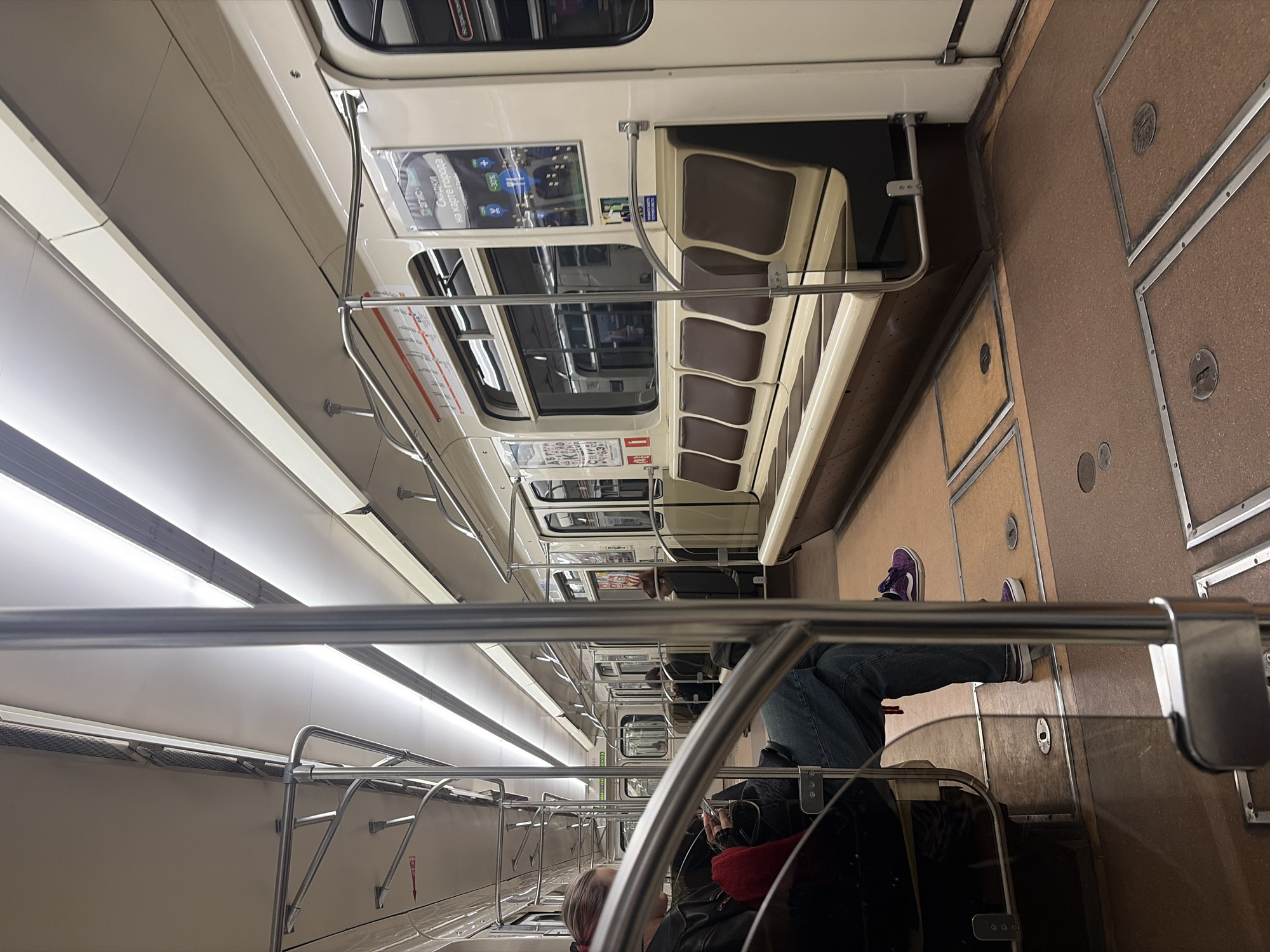 The image captures the interior of a subway car, likely in Saint Petersburg, Russia, based on the architectural style and design of the train. Passengers are visible in the car, some seated, others standing. The lighting is bright and artificial. The car features rows of seats with brown and beige upholstery, as well as metal handrails for standing passengers. The walls of the car are off-white, with brown panels along the floor. A rider has placed his legs over onto the seats across from him. The scene portrays a typical commute on a public transportation system.