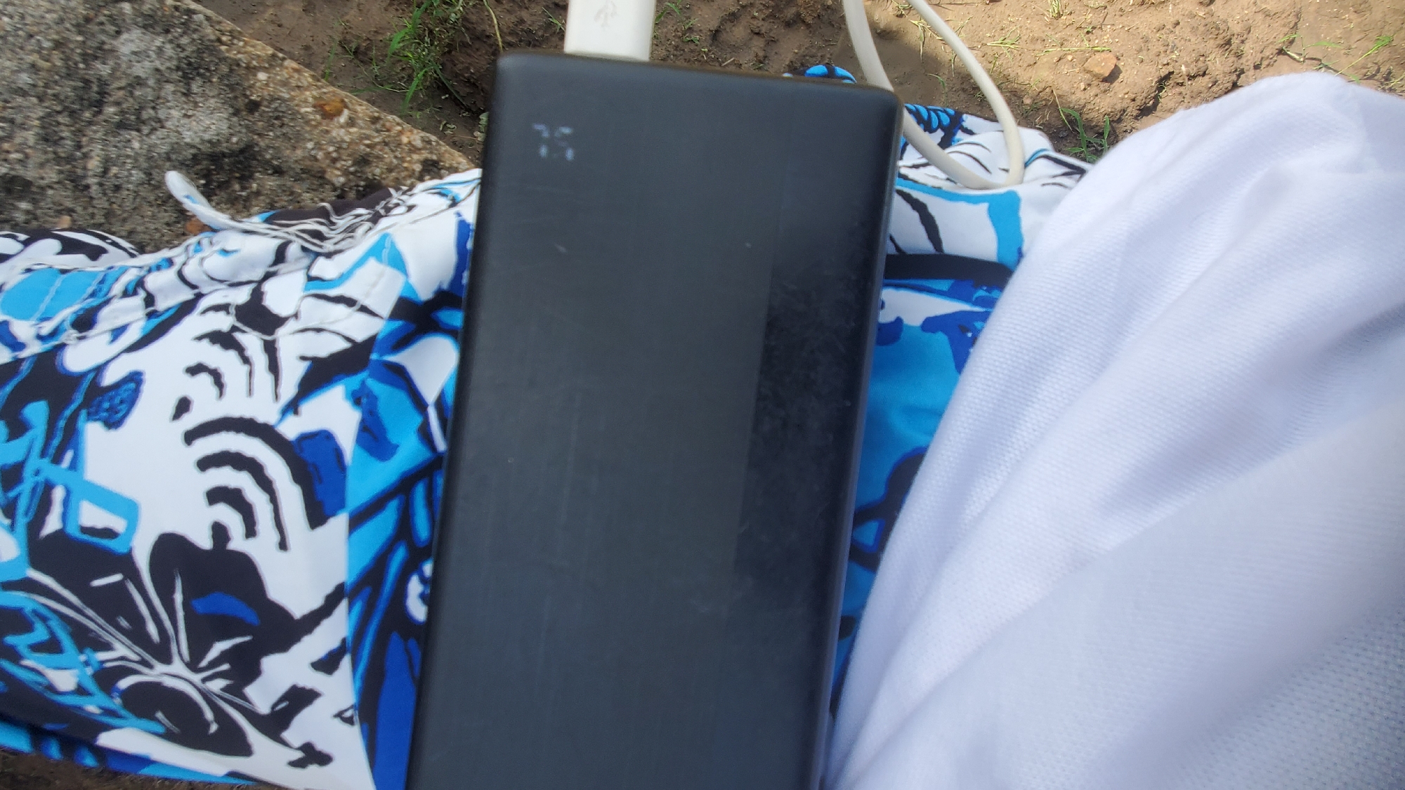 The image shows a black rectangular power bank resting on a person's lap, which is covered by a blue and white patterned fabric. A white charging cable is plugged into the power bank and extends out of frame. The background appears to be an outdoor setting with dirt and grass. The power bank has a digital display showing "75". The perspective is a close-up shot, with the power bank occupying the majority of the frame.