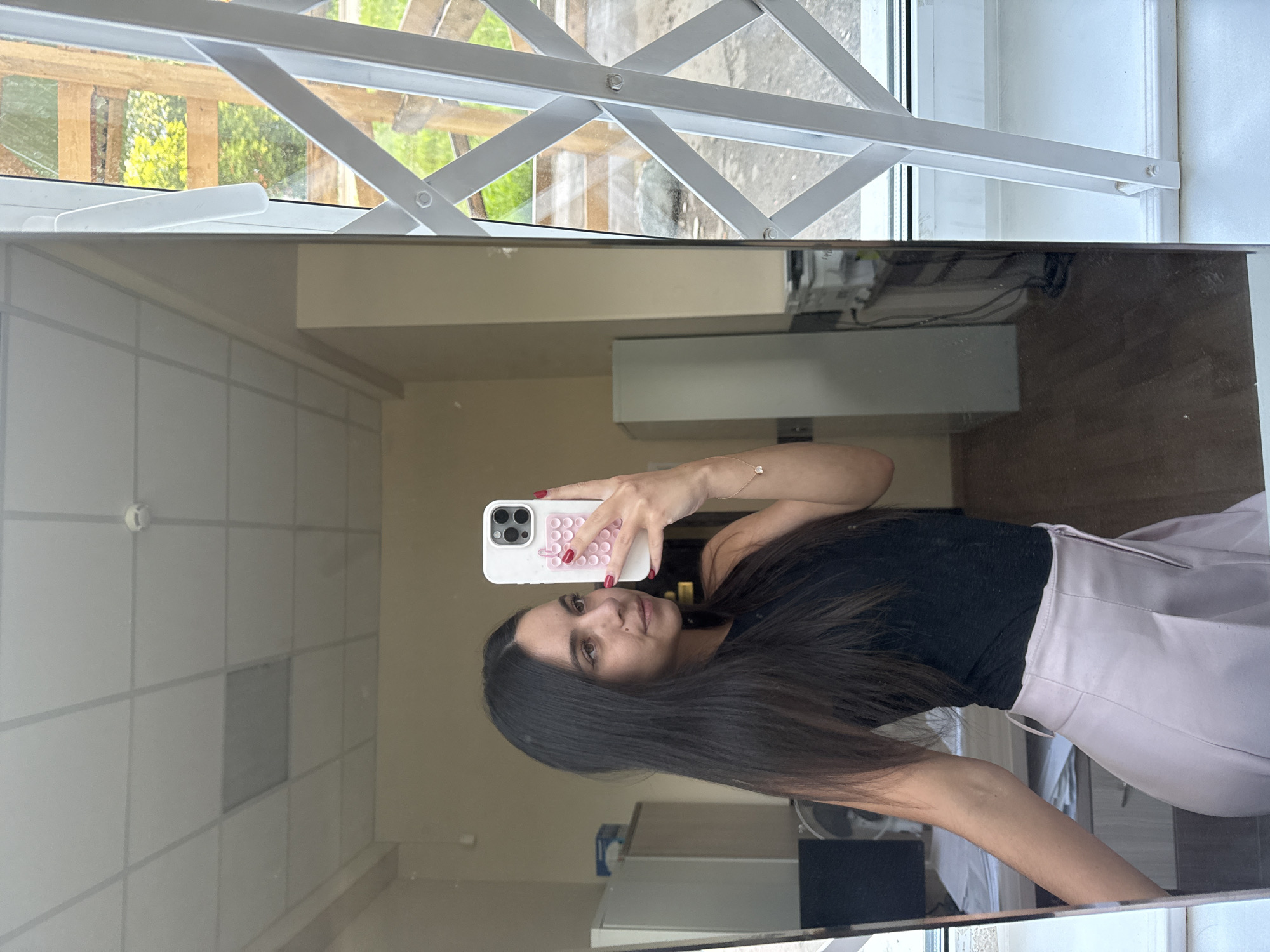 In an indoor setting, a young woman with long, dark hair takes a selfie in a full-length mirror. She is wearing a black tank top and a light pink pleated skirt. Her right hand, holding a white smartphone, is visible in the reflection. The background of the room shows filing cabinets, a desk with a computer monitor, and a window with a metal security grate. The ceiling appears to be a drop ceiling with fluorescent lighting.