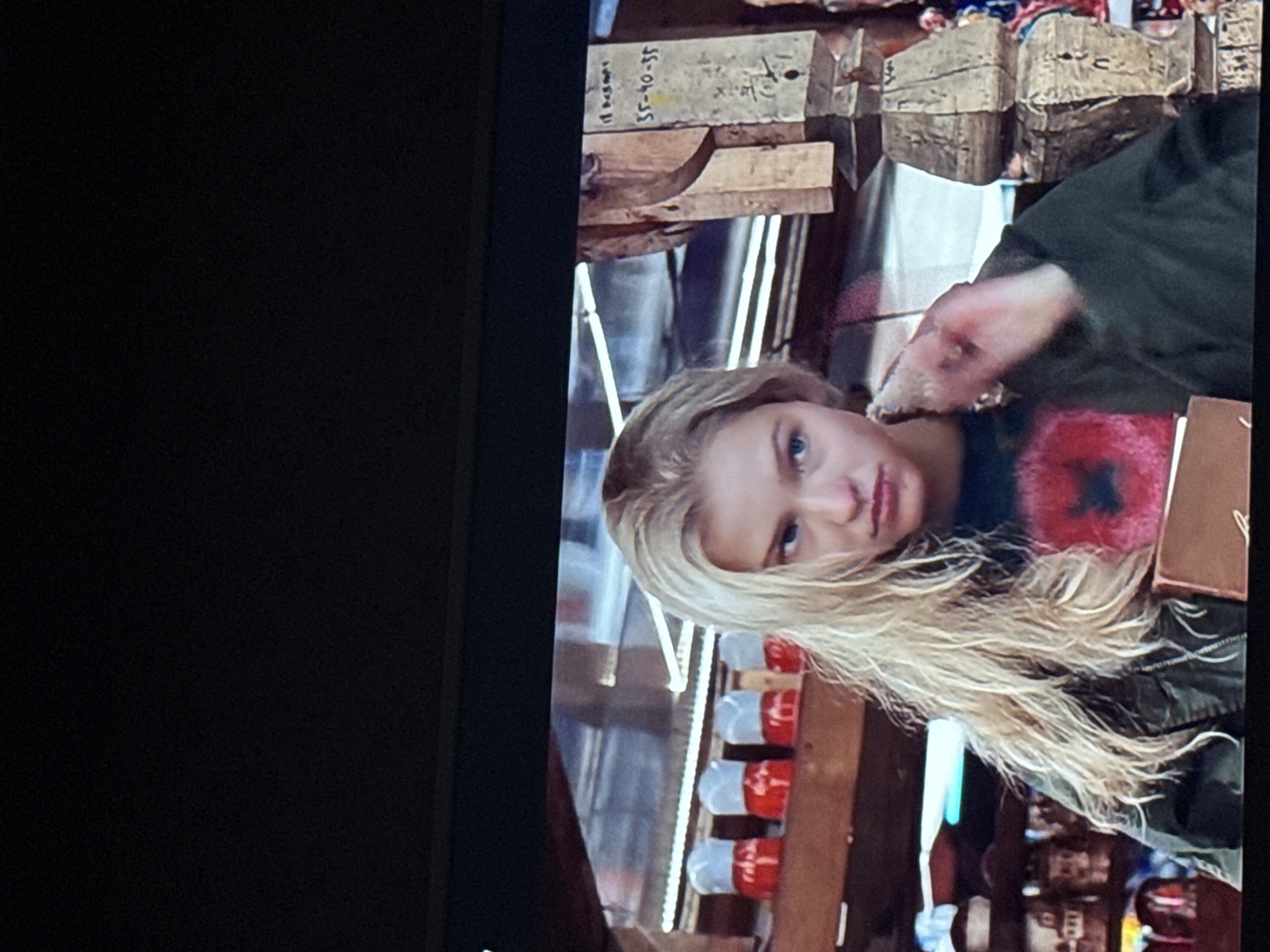 A woman with blonde hair is visible. She is wearing a dark green jacket. Behind her, there are shelves with what appear to be goods for sale, including items that look like large pills. There is also wooden framing and other objects on the shelves. Writing is visible on some of the items in the background, including the numbers "35-40-35". The image is taken from an angle, and part of the left side of the image is black. The location cannot be determined from the image.