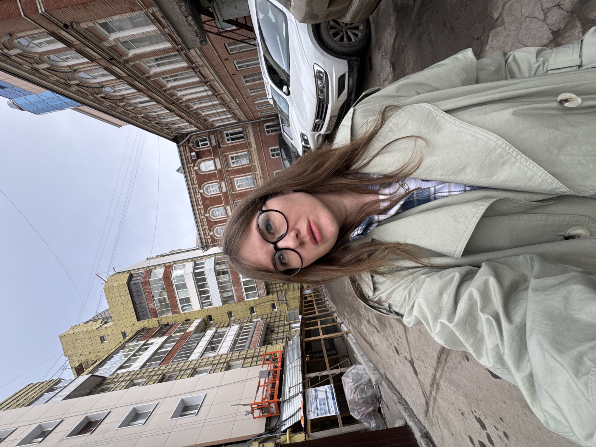 The image depicts a female taking a selfie on a city street in Perm, Russia, specifically identified as Petropavlovskaya Street, Building 46. The woman, appearing to be in her late twenties to early thirties, has long, light brown hair and wears black-framed circular glasses, a light beige or pale green trench coat, and a blue and white plaid shirt. She looks directly into the camera with a neutral expression.
The scene is set outdoors under an overcast sky. To the immediate left, a multi-story building, likely residential, is undergoing renovation; its facade is partially covered with yellow insulation material and scaffolding. A large orange scissor lift is positioned on a temporary platform near the building. Below this construction, a temporary sheltered walkway with a metal roof is visible, along with a white sign containing Cyrillic text that explicitly states "Г. ПЕРМЬ, УЛ. ПЕТРОПАВЛОВСКАЯ, Д. 46."
On the right side of the frame, an older, multi-story building with reddish-brown brickwork and numerous arched windows with white frames stands prominently. Above this historic structure, a modern building with a blue, reflective glass facade is partially visible. Several vehicles, including at least two white SUVs, are parked along the curb on the right, one of which is a Volkswagen Tiguan with a partially visible Russian license plate showing "179 VO 59." The ground is a cracked, dark paved surface, possibly asphalt or concrete, showing signs of wear and some minor pooling or dampness. Overhead, multiple thin wires traverse the sky between the buildings.