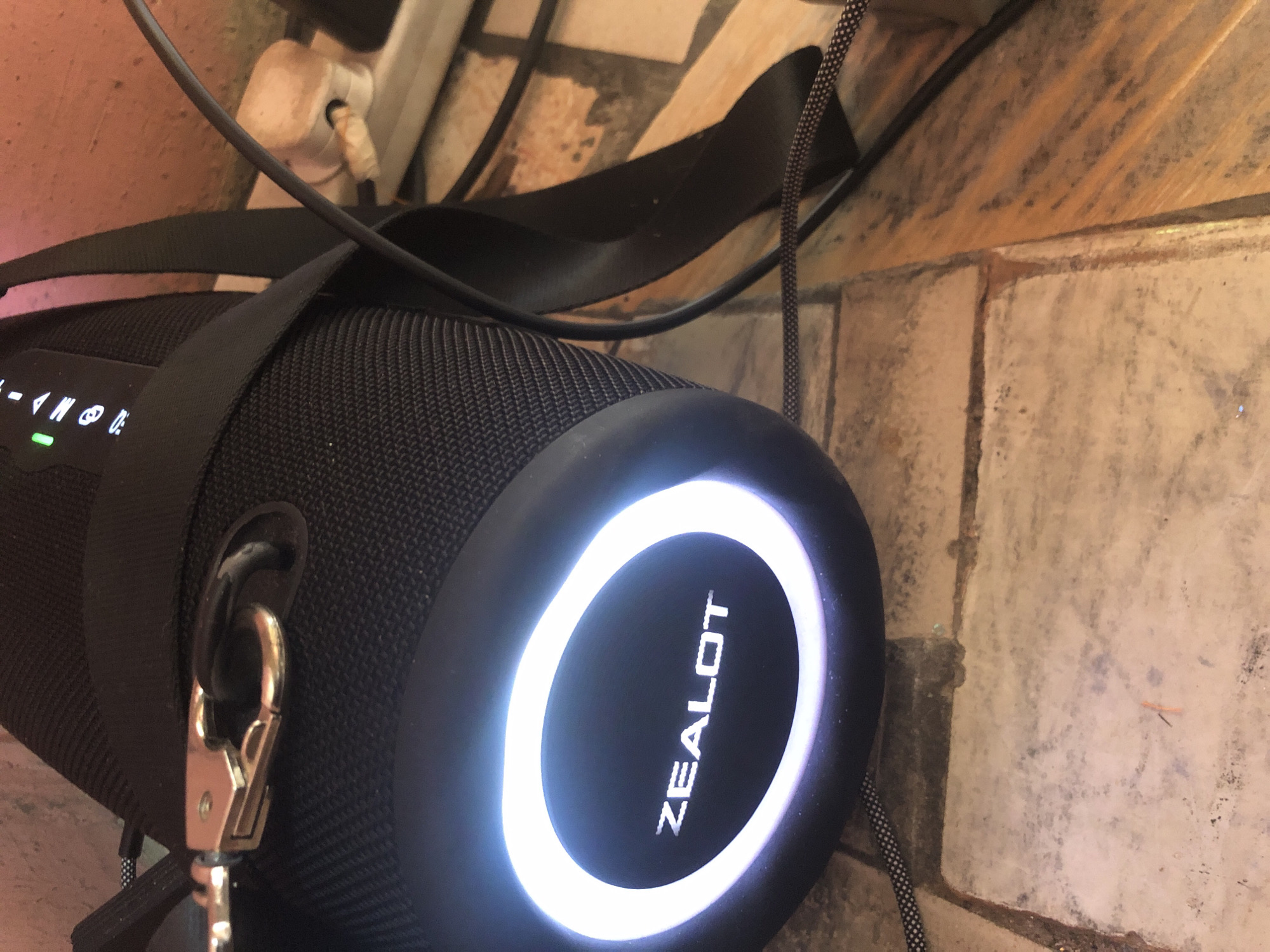 In Ado, Nigeria, a black portable speaker with a light-up "ZEALOT" logo sits on a tiled floor. A black strap is attached to the speaker, with a metal clasp visible. A power cord snakes away from the speaker and plugs into a white power strip, which has a white electrical adapter plugged into it. The background consists of a textured wall.