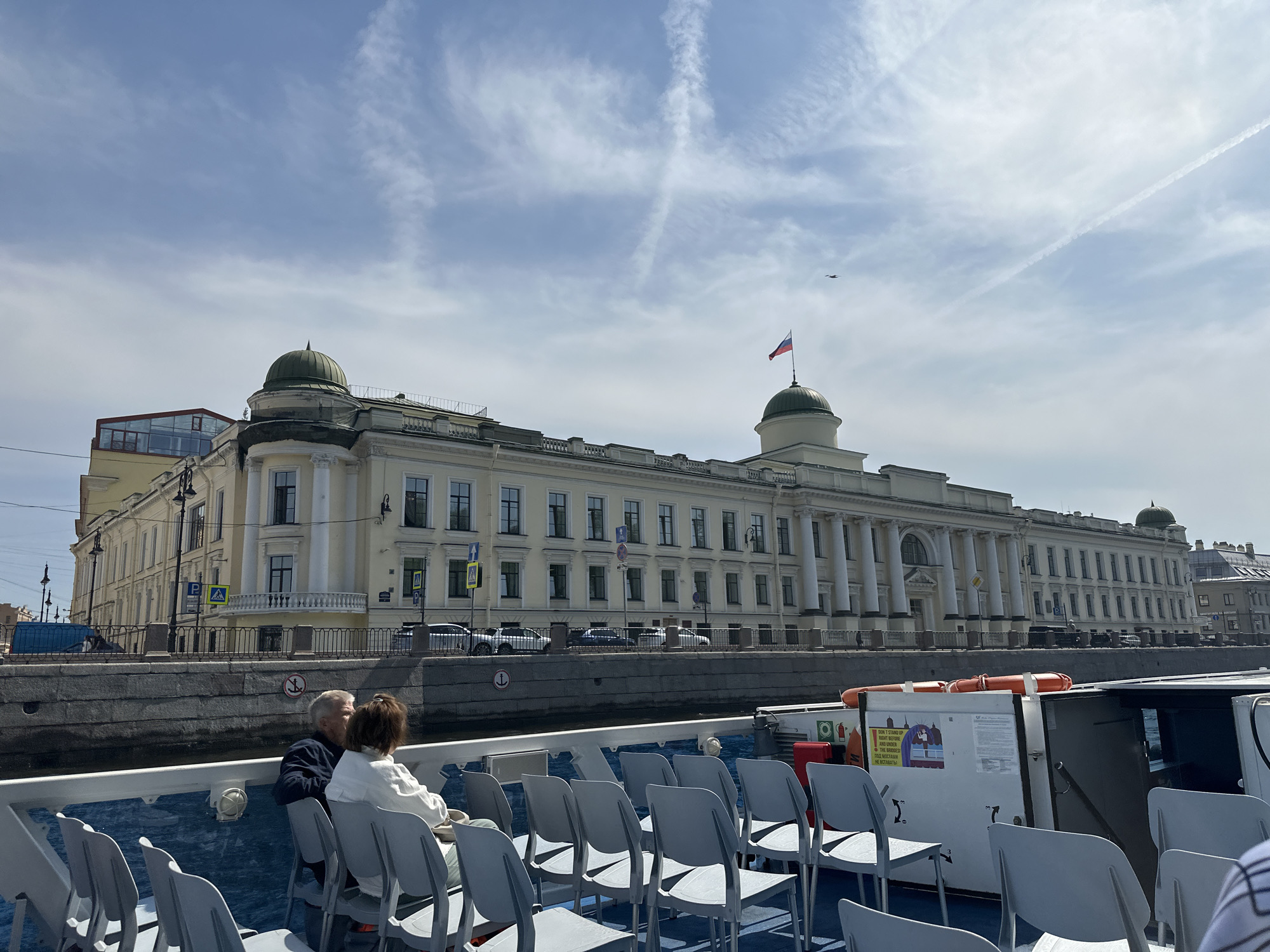 The image shows a view from a boat on a canal in Saint Petersburg, Russia. On the bank, there is a long, light-yellow building with a green dome, classical columns, and the Russian flag on top. Several cars are parked along the street in front of the building. On the boat, rows of grey plastic chairs are visible. Two people, an older man and woman, are seated near the front of the boat. The sky is blue with streaks of white clouds. An airplane is also visible in the sky.