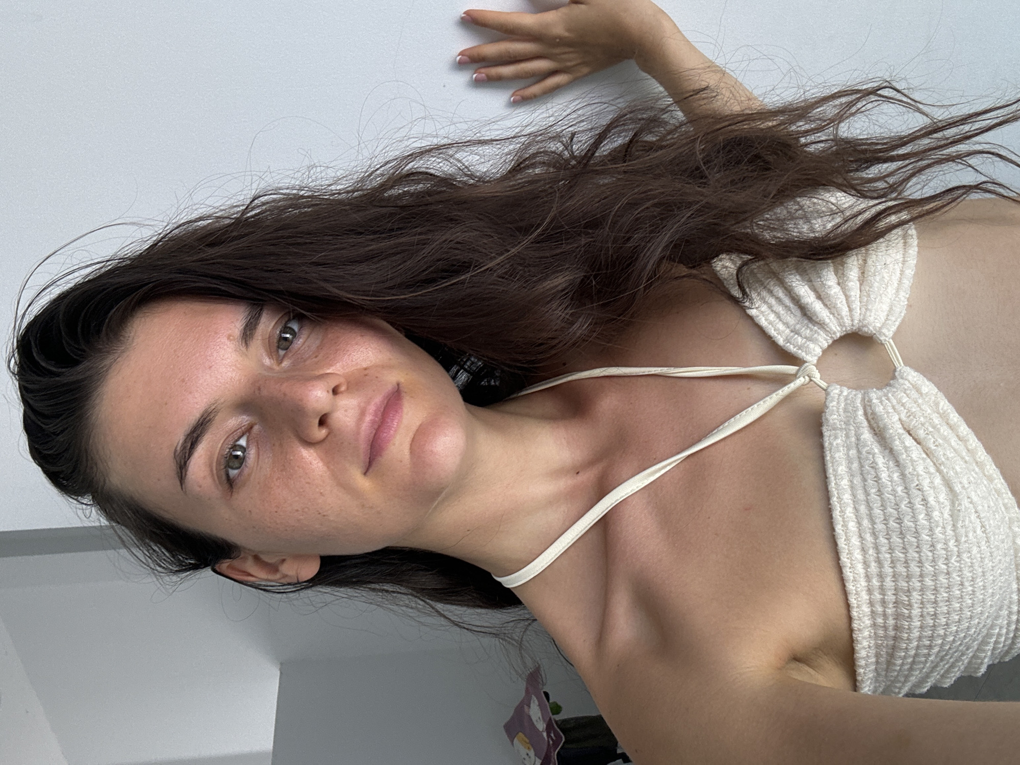 A woman with long, dark brown hair and fair skin is pictured from a low-angle point of view. She is wearing a white, knit top with thin straps. Her left hand is extended and resting against a plain white wall. The background appears to be a simple, indoor setting, likely a room with white walls and a glimpse of some items in the lower corner. No specific elements within the image definitively place the scene in Mũi Né, Vietnam.