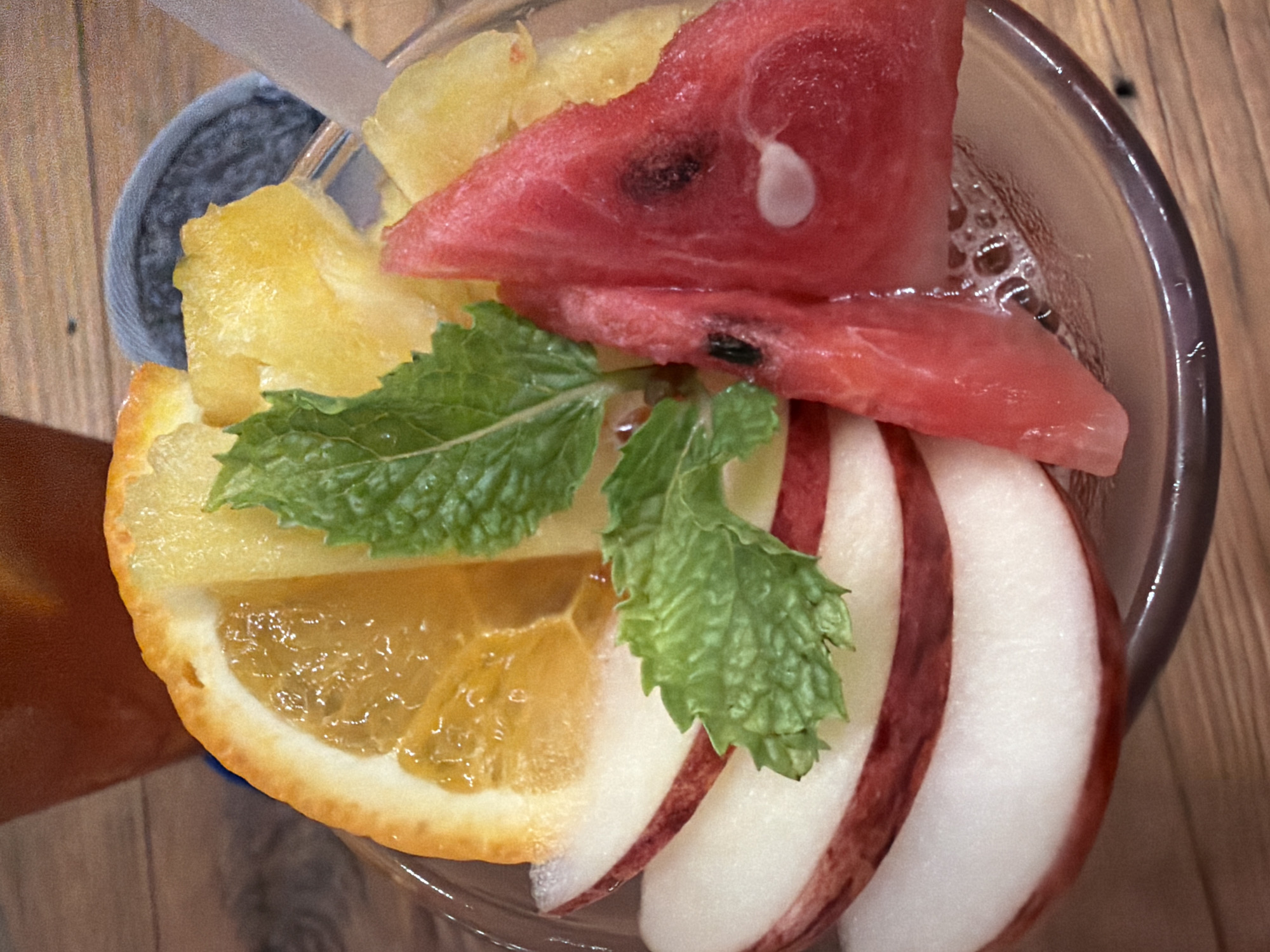 The image shows a close-up view of a glass filled with fruit and ice, possibly a beverage. The glass is on a wooden surface. Inside the glass, there are slices of orange, apple, watermelon, and pineapple. A sprig of mint is also visible, adding a touch of green to the colorful arrangement. The background is slightly blurred, but appears to be another glass or container with a dark liquid and a wooden surface, suggesting a cafe or restaurant setting. The lighting suggests an indoor environment. No people or specific location markers are present in the image.
