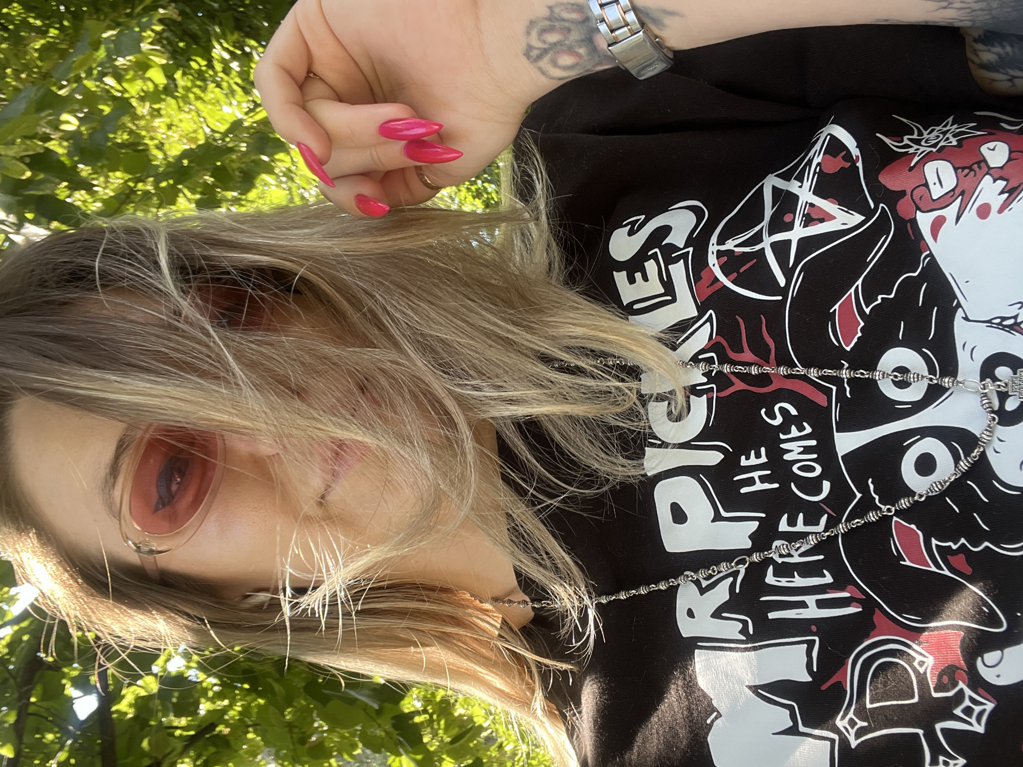 The image is a close-up, low-angle shot of a person outdoors, with a lush green canopy of leaves framing the top and bottom of the frame, suggesting a sunny day. The person has blonde hair, some of which falls over their face, and is wearing pink-tinted sunglasses. They are also wearing a black t-shirt with a graphic print. On the shirt, the words "HE HERE COMES" are visible in white text. The graphic also includes a pentagram symbol and cartoonish figures. The person's left hand is visible in the upper portion of the frame, with bright pink, pointed fingernails. They are wearing a silver watch on their left wrist, and a silver chain necklace is visible on their chest. The lighting suggests it is daytime, and the presence of leaves indicates an outdoor setting, possibly a park or a garden. There are no explicit emotional cues visible due to the sunglasses and framing.