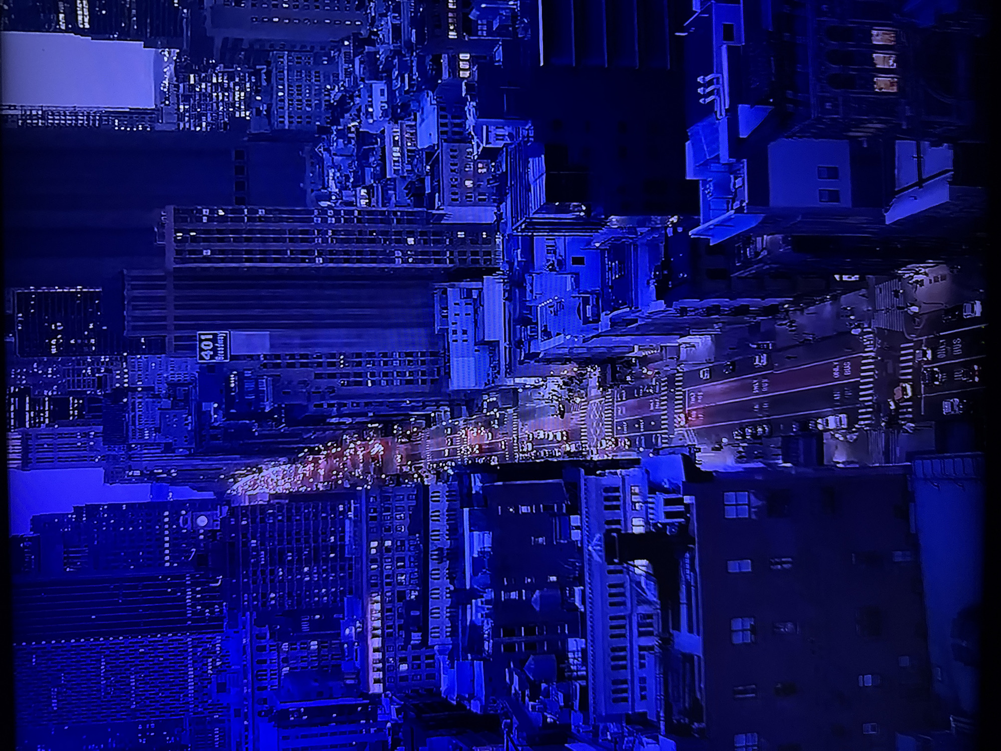 The image displays an aerial, nighttime view of a busy city street, illuminated by artificial lights. Tall buildings, characteristic of a dense urban environment, line both sides of the street, their windows lit with a warm glow. Cars are visible on the street, their headlights and taillights creating streaks of red and white light, indicating constant traffic flow. Pedestrians can be seen on the sidewalks, adding to the sense of activity and life. The overall color palette is dominated by deep blues and the warm tones of the lights, suggesting either twilight or nighttime. The scene captures the vibrant energy of a metropolis after dark.
The location of Ruse, Bulgaria, is not depicted in this image. This scene is representative of a major urban center, likely in North America or Europe, with its high-rise architecture and bustling street life.