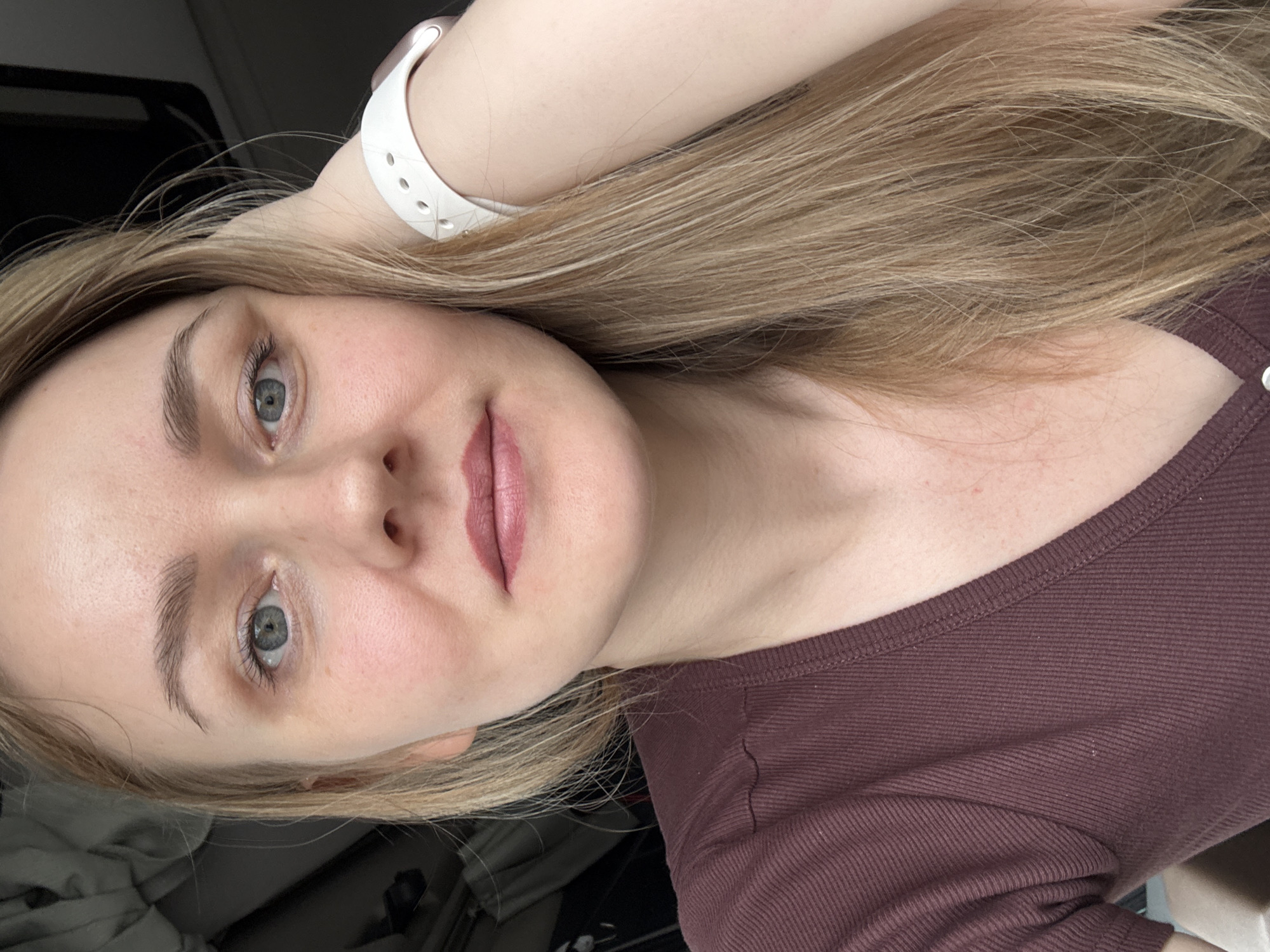 A close-up, selfie-style shot of a fair-skinned woman with blonde hair. Her right arm is raised near her head, a white smartwatch visible on her wrist. She is wearing a maroon-colored, crew-neck top. Her face is visible, with prominent eyebrows, grey eyes, and lips painted in a mauve shade. The background is out of focus, appearing to be indoors, potentially near a car. There are no indications this photo was taken in Kukui, Russia.