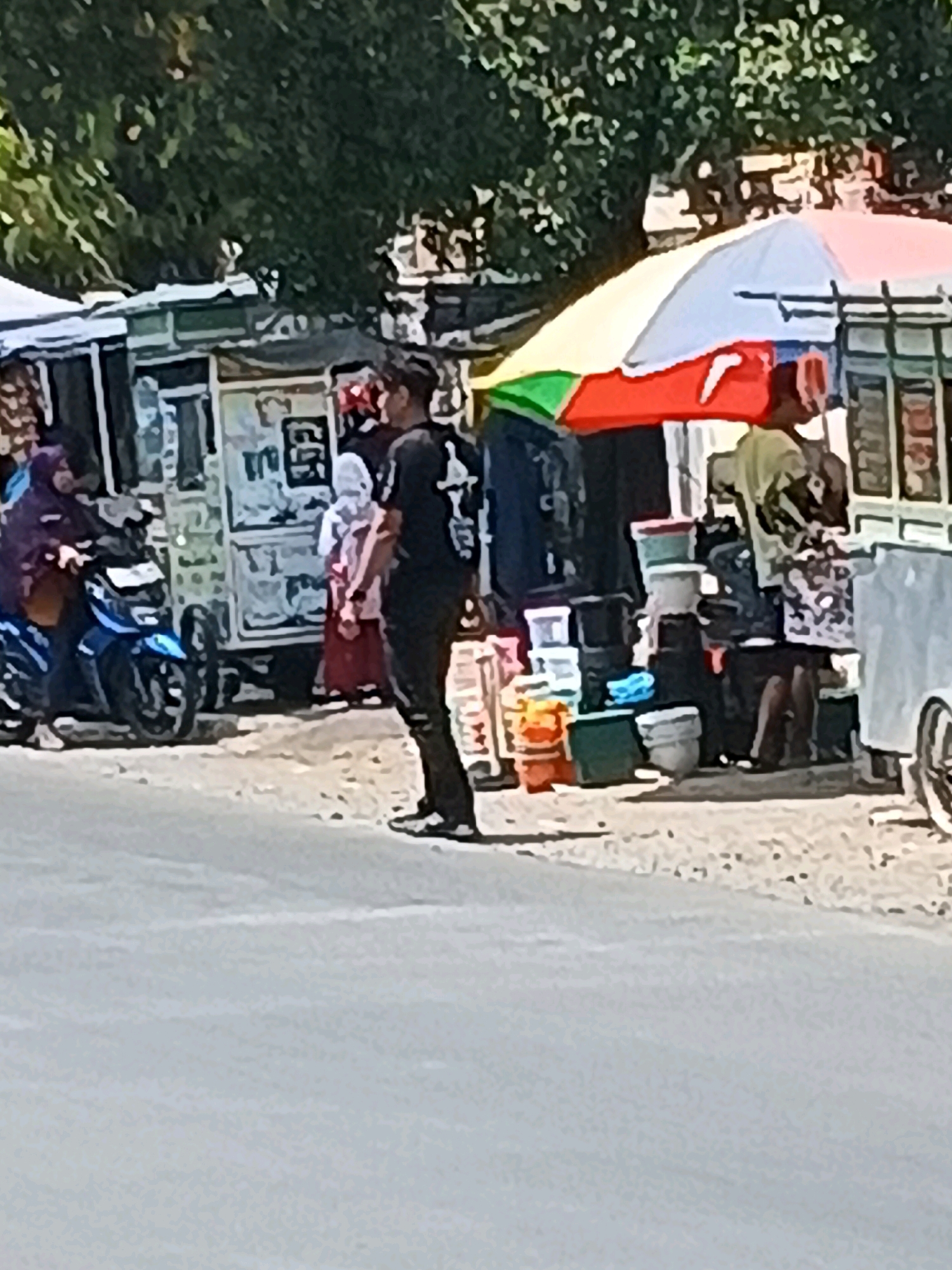 The image depicts a street scene in Taman, Indonesia, with several market stalls and people. A person is standing on the right side of the image, facing away from the camera, wearing a black t-shirt and black pants. To their left, a woman in a purple hijab is riding a blue motorcycle. Behind the woman and motorcycle, there are market stalls with various items displayed. A colorful umbrella shades some of the stalls. Several containers, possibly for food or goods, are stacked near the stalls. The ground appears to be paved, and there are trees in the background. The overall scene suggests a bustling local market on a sunny day.