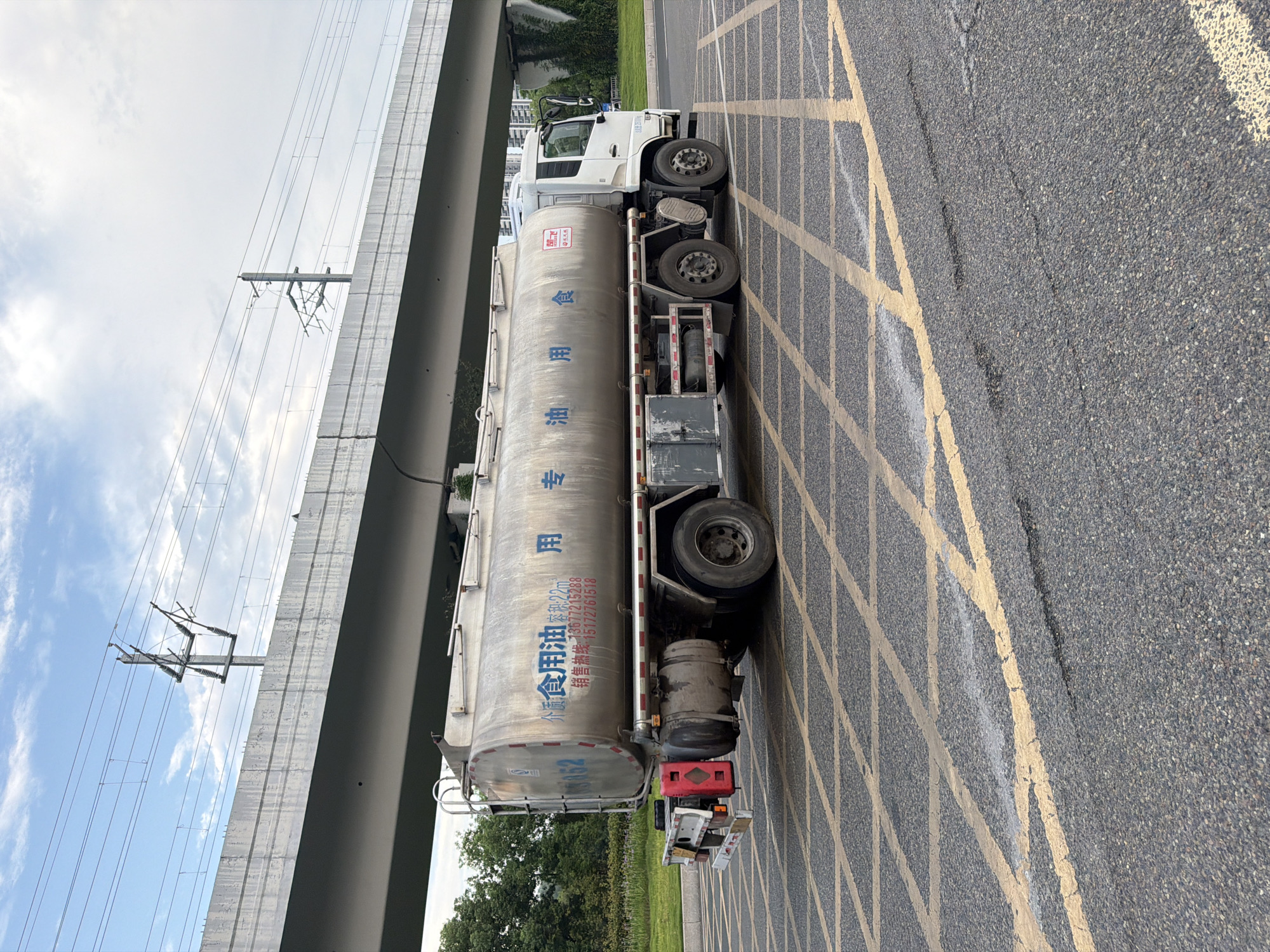 A white tanker truck with a large silver cylindrical tank is parked on a city street in Jiangjin, China. The truck has Chinese characters on the side of the tank, which appear to be related to edible oil. The truck is parked on an asphalt road marked with a yellow grid pattern. In the background, an elevated concrete structure, likely a highway or railway overpass, spans across the scene, with power lines and poles visible above it. Green trees and vegetation line the side of the road, and modern buildings can be seen in the distance. The sky is partly cloudy.