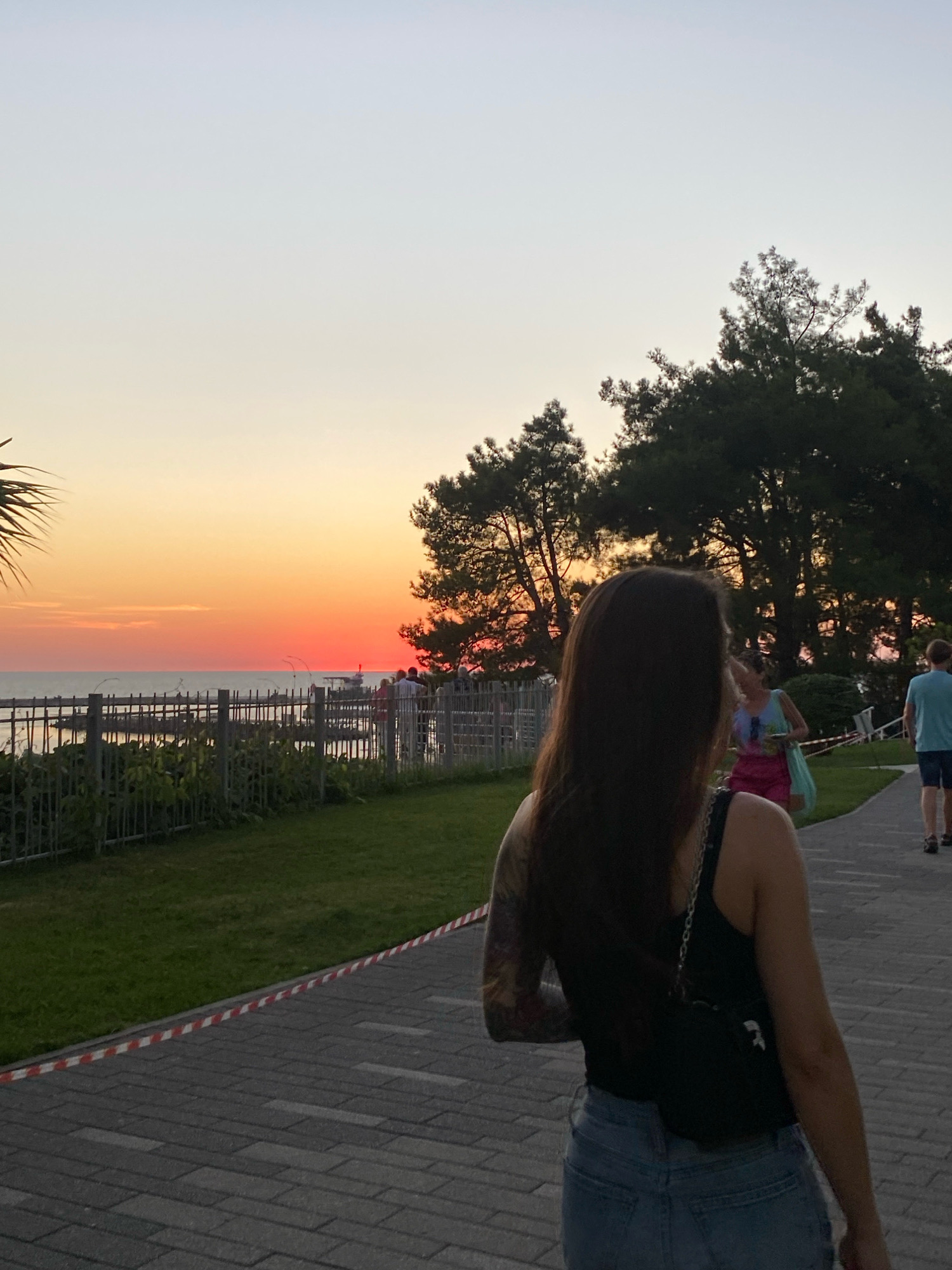 The image captures a sunset scene along the Black Sea coast in Sochi, Russia. In the foreground, a woman with long, dark hair and a tattoo on her left arm walks away from the camera on a paved walkway. She is wearing a black tank top and denim shorts, carrying a small black bag with a chain strap. The walkway is lined with grass and a fence separating it from the sea. Beyond the fence, the ocean stretches out, with a vibrant orange and red sunset casting a glow over the water. Several people are visible in the distance, some walking along the promenade, and a structure resembling a pier or jetty extends into the sea. Tall, dark green trees line the edge of the walkway, creating a silhouette against the colorful sky. The overall atmosphere is serene, with the warm hues of the sunset dominating the scene.