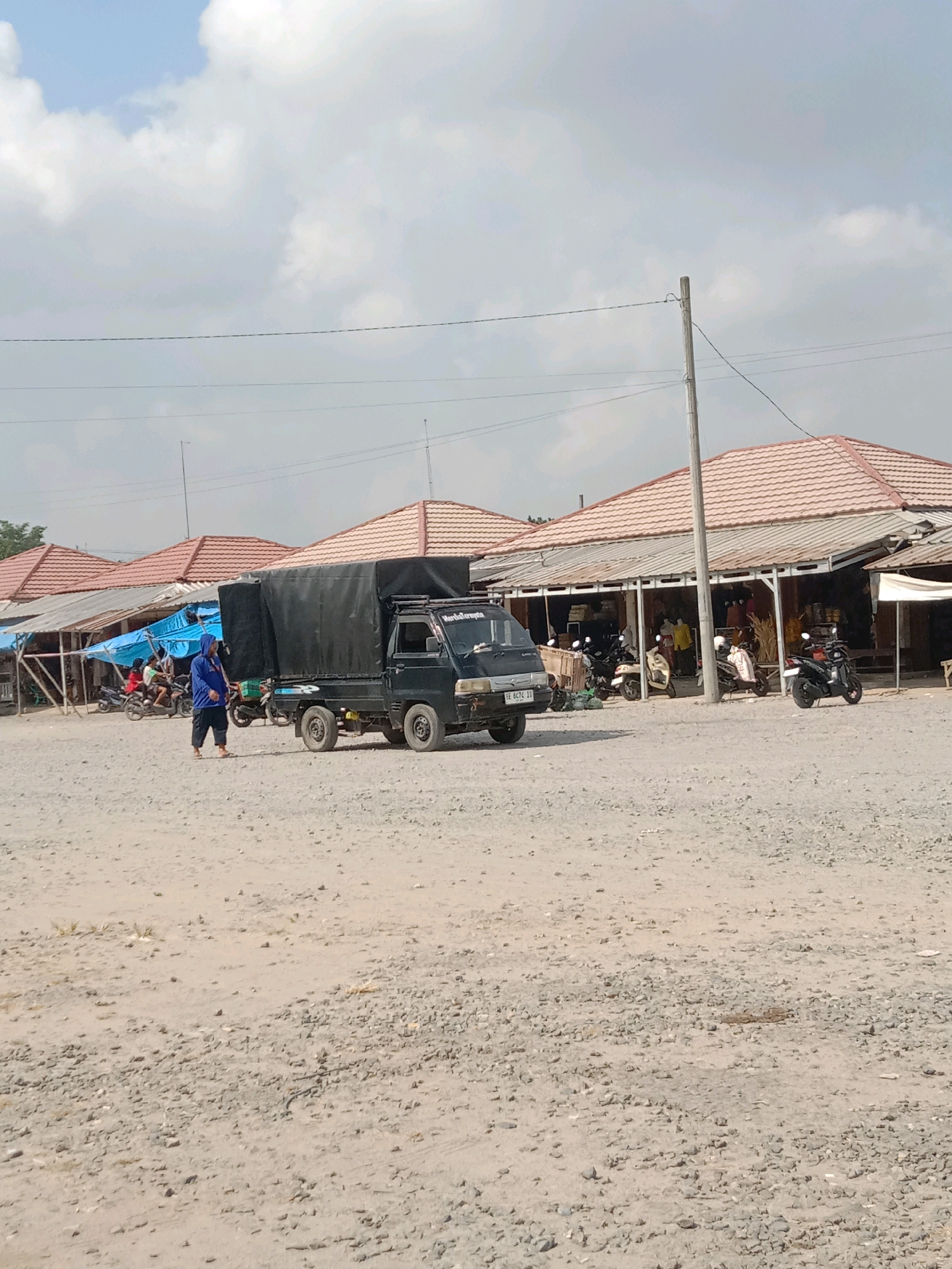 The image depicts an outdoor market scene with a gravel and dirt ground. In the foreground, a person in a blue hooded jacket and dark pants is walking towards the right. To their left, a black truck with a black tarp covering its cargo bed is parked. The truck has the license plate "BE 6474 IV". Behind the truck, a row of market stalls with tiled roofs and open fronts are visible. Several motorcycles are parked in front of the stalls. Some people can be seen inside or in front of the stalls. The sky is cloudy. The location is identified as the city of Fajar Mataram, Indonesia.
